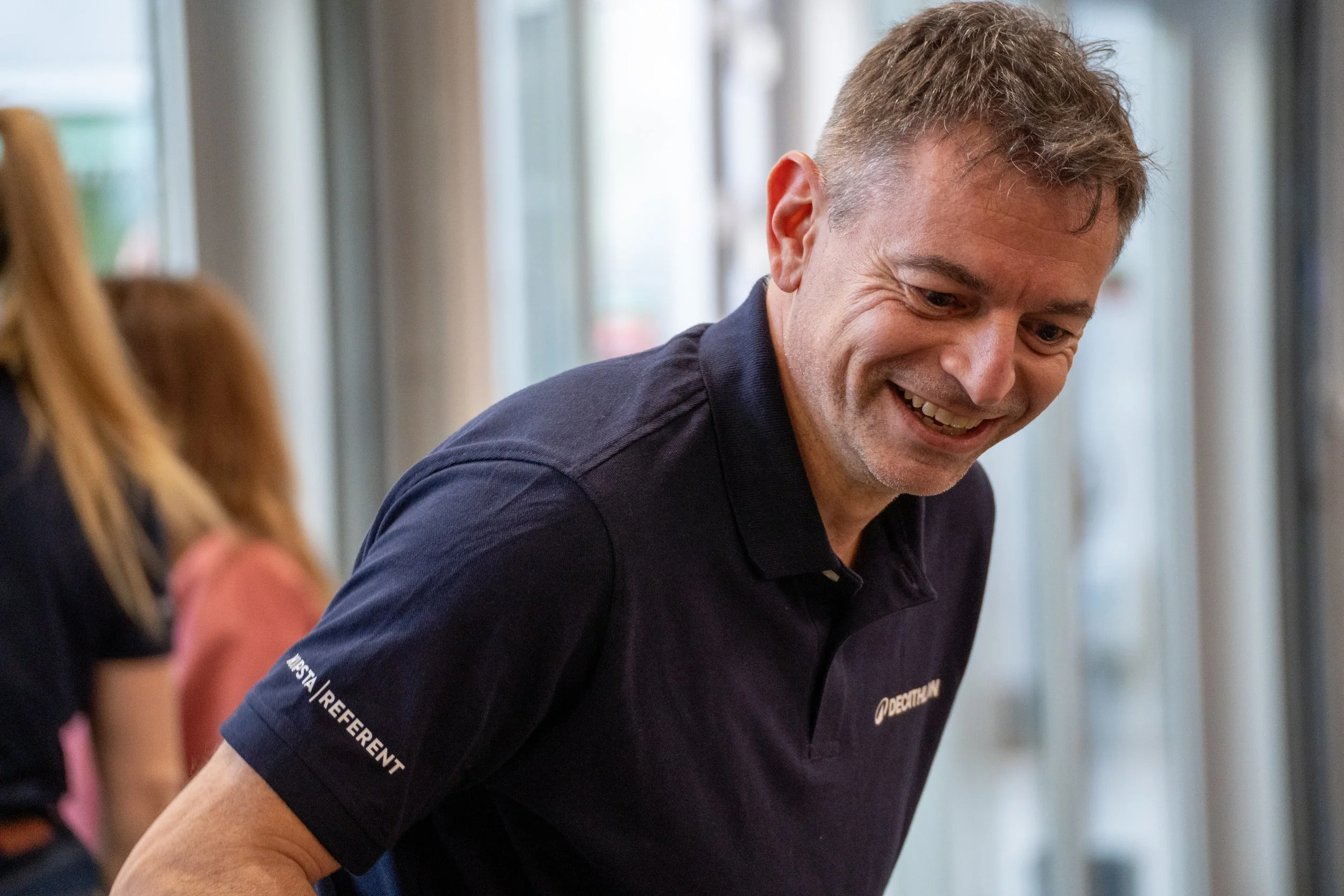 Un homme souriant portant un polo noir avec le logo Decathlon, dans un environnement intérieur lumineux.