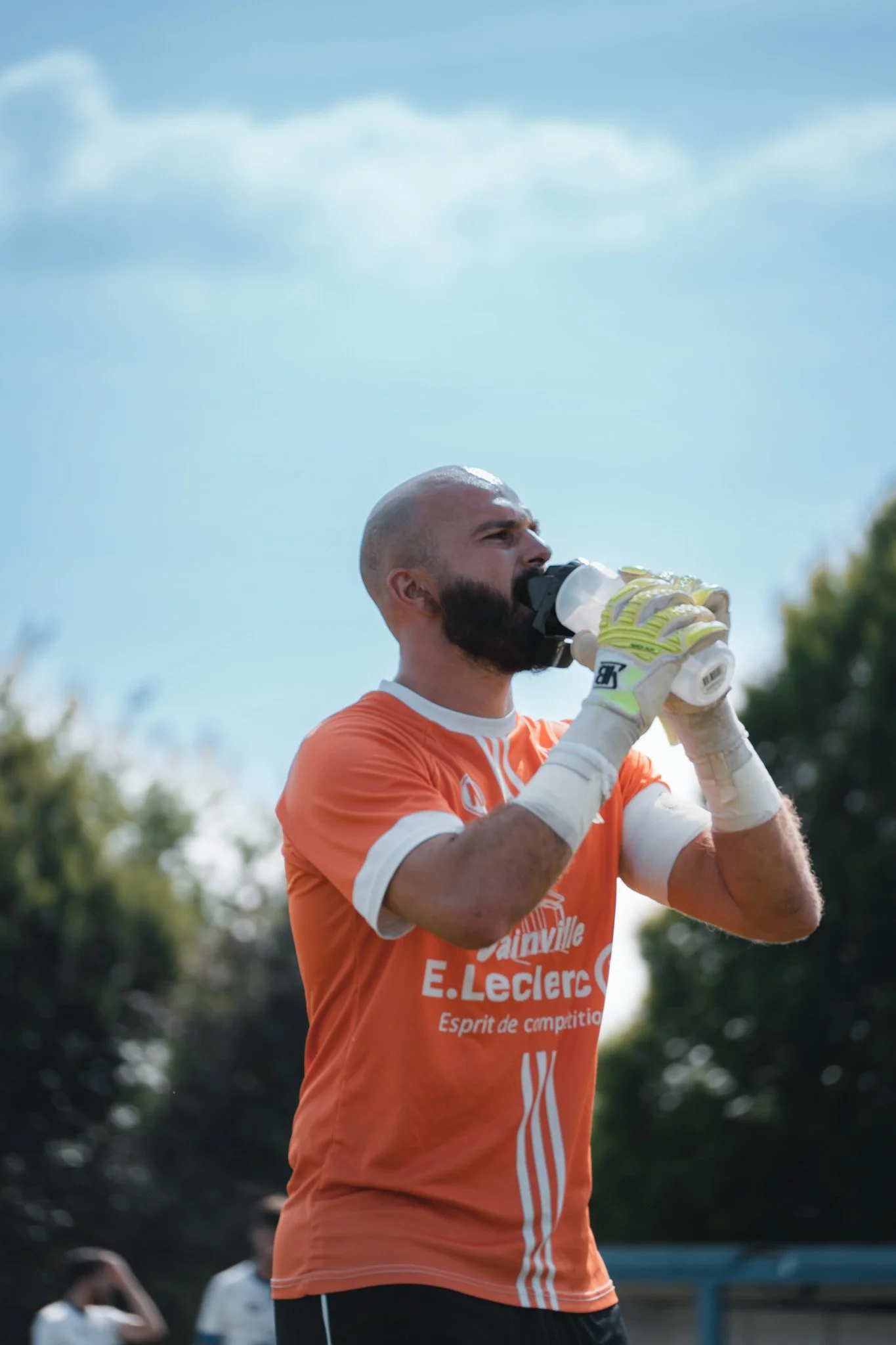 Un joueur de football en tenue orange boit de l'eau à la mi-temps ou après un match, en extérieur.