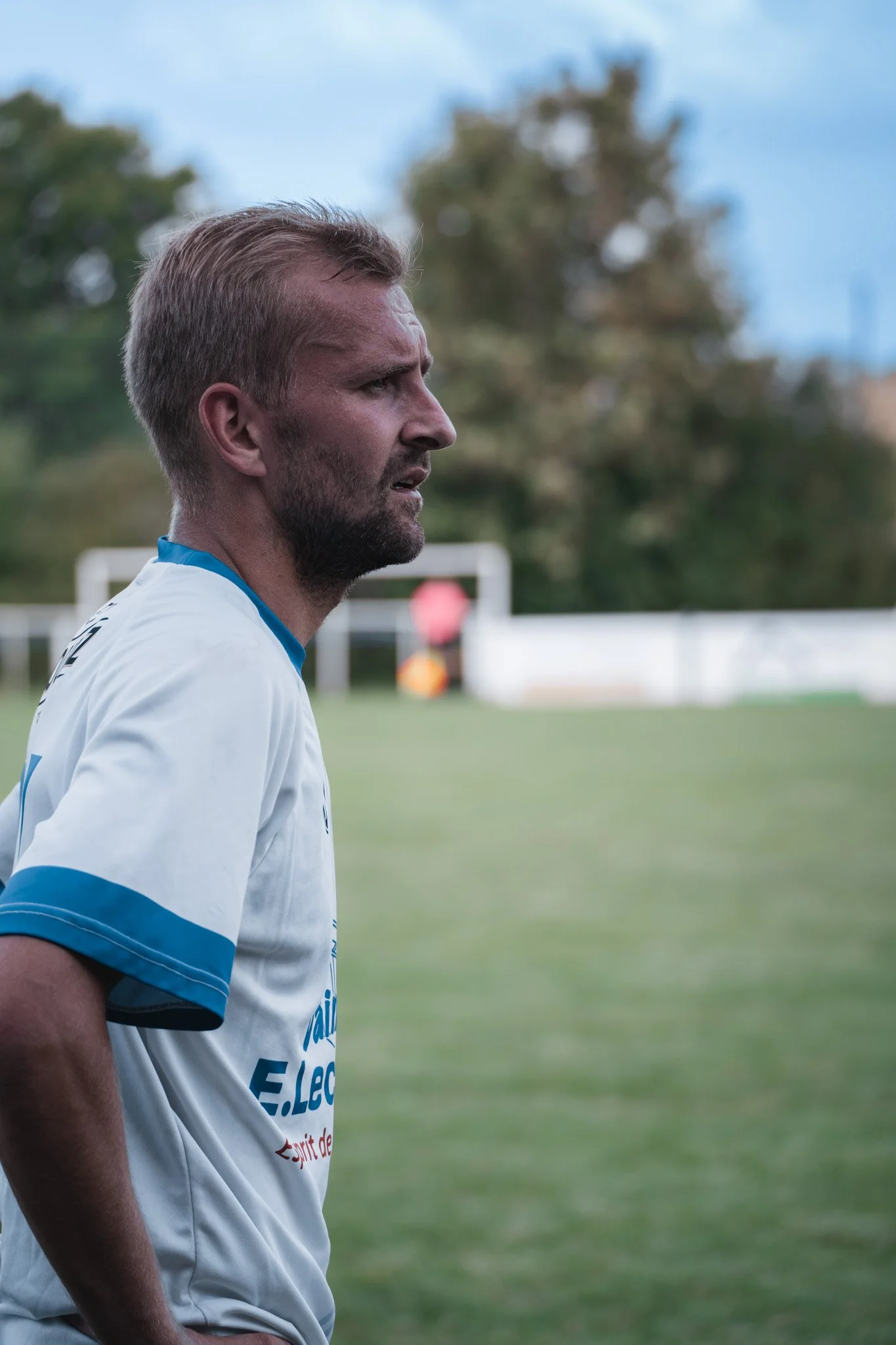 Un homme en maillot de football regarde vers la droite sur un terrain de football en extérieur.
