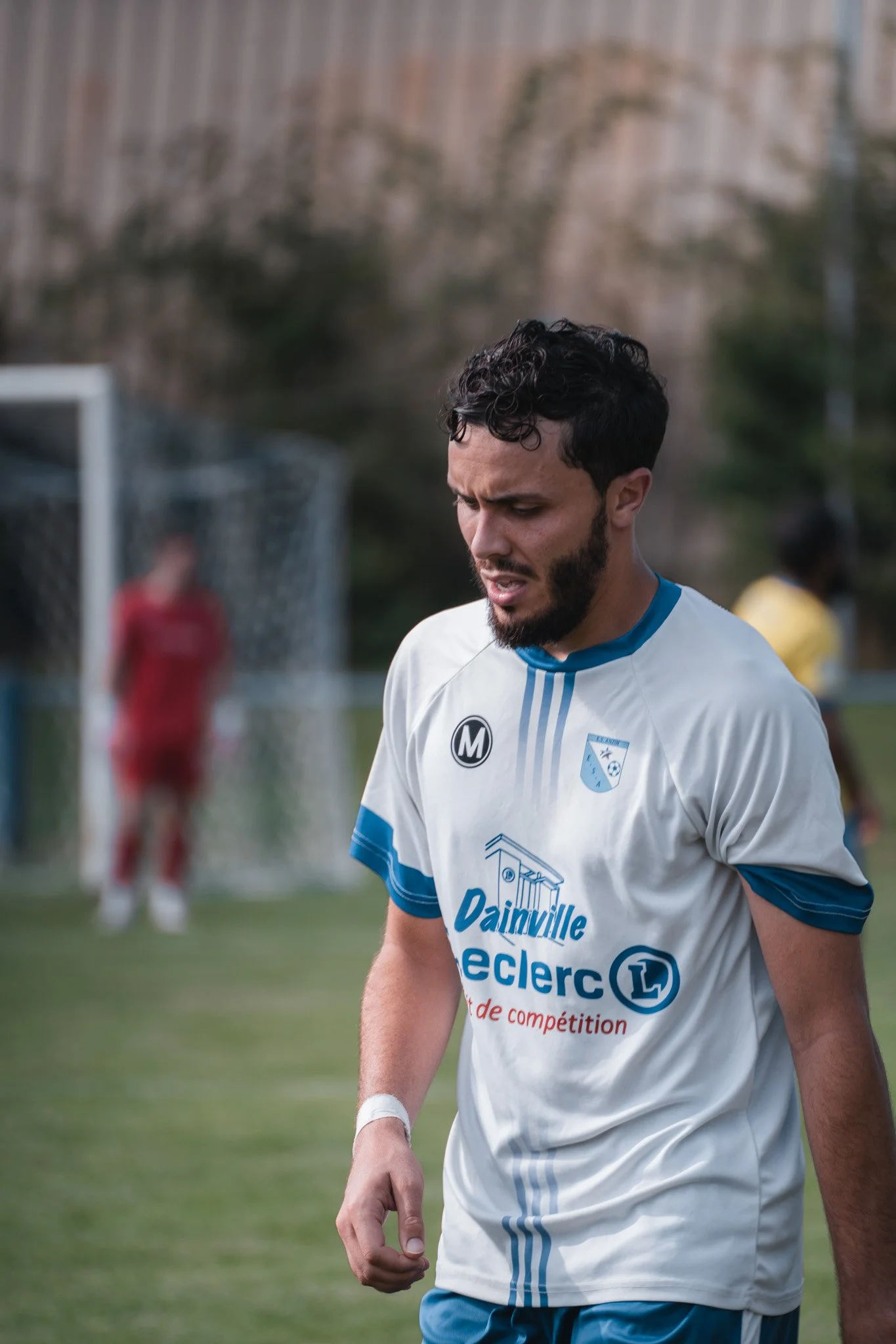 Un joueur de football en maillot blanc avec des accents bleus, casque et maillot avec logos d'une équipe, semble déçu ou fatigué, sur un terrain de football avec un joueur en costume rouge et un autre en jaune au fond.