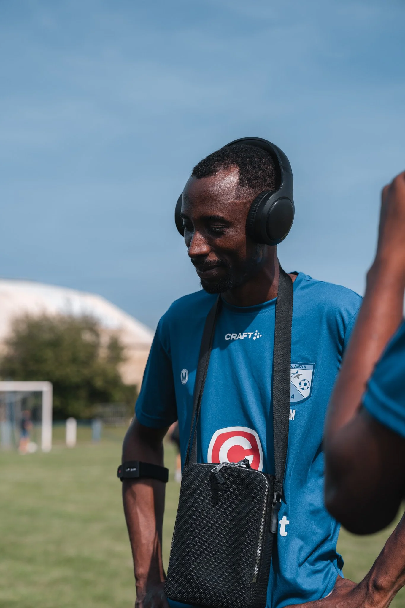 Homme portant un t-shirt de soccer, un casque audio et une sacoche en bandoulière, dans un terrain de sport outdoor.