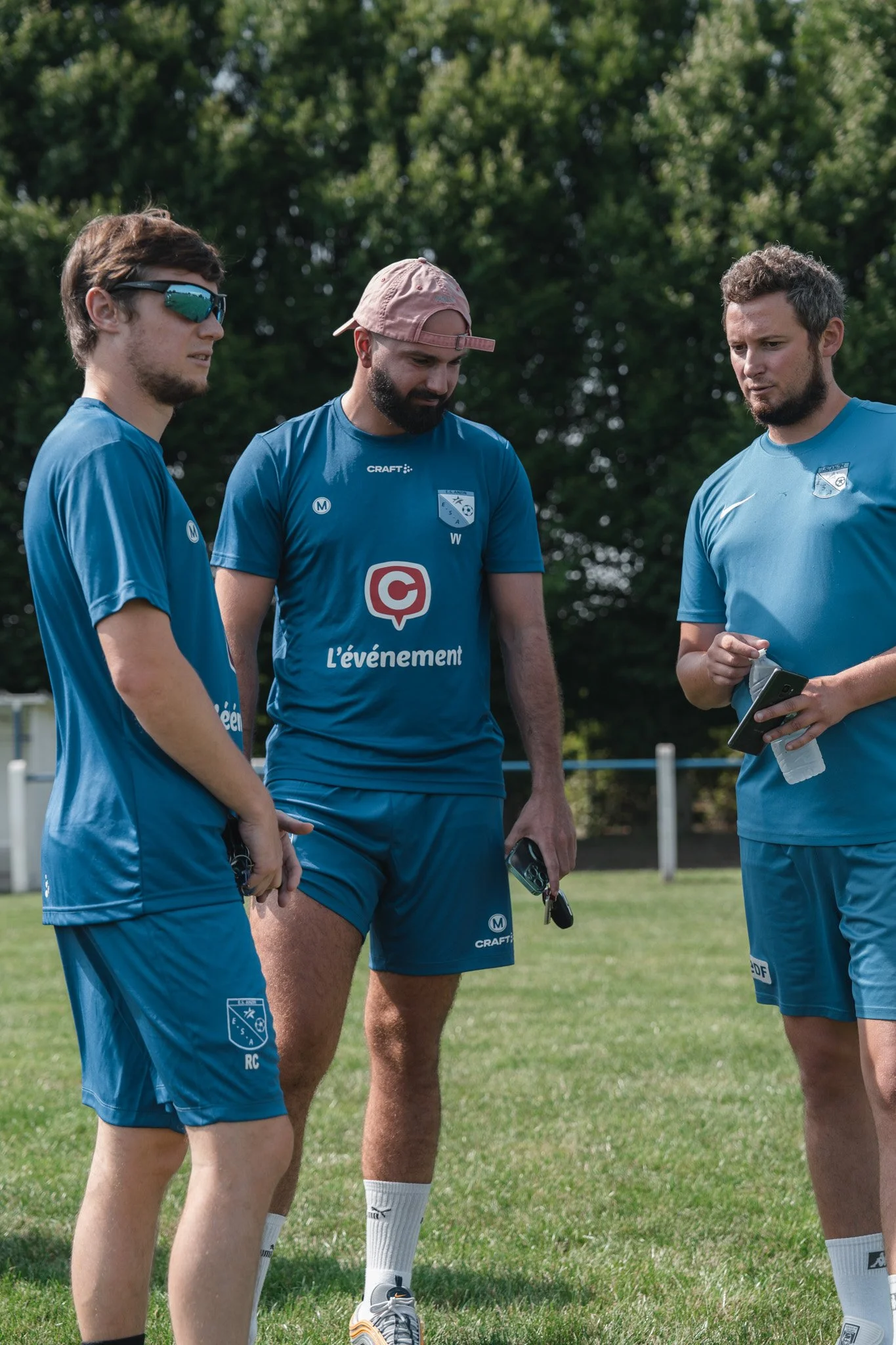 Trois hommes en tenue de sport bleue discutent sur un terrain herbeux avec des arbres en arrière-plan, en pleine journée.
