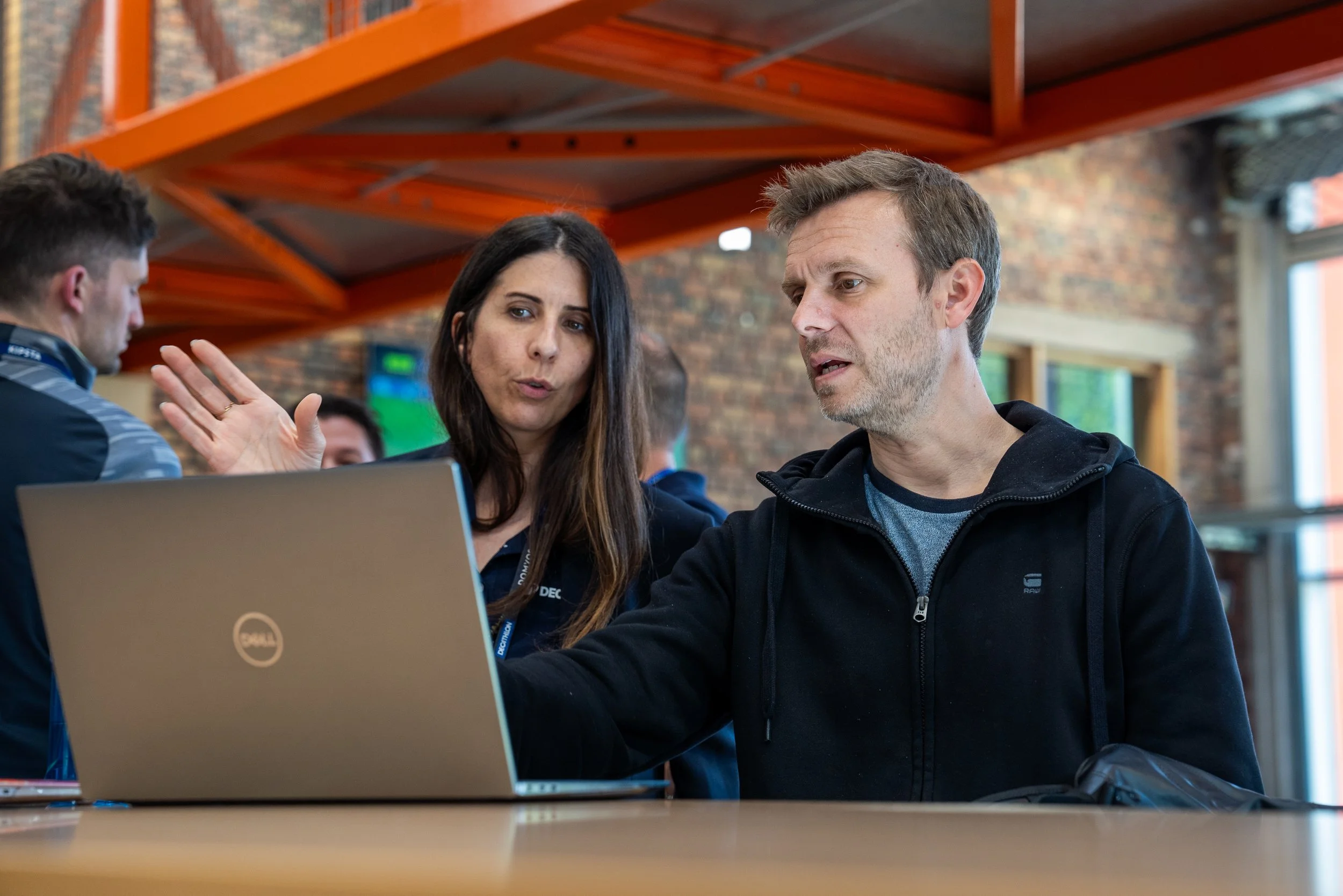 Deux personnes discutent devant un ordinateur portable Dell dans un espace intérieur avec un plafond orange et des murs en briques. Une femme parle, tandis qu'un homme écoute attentivement.