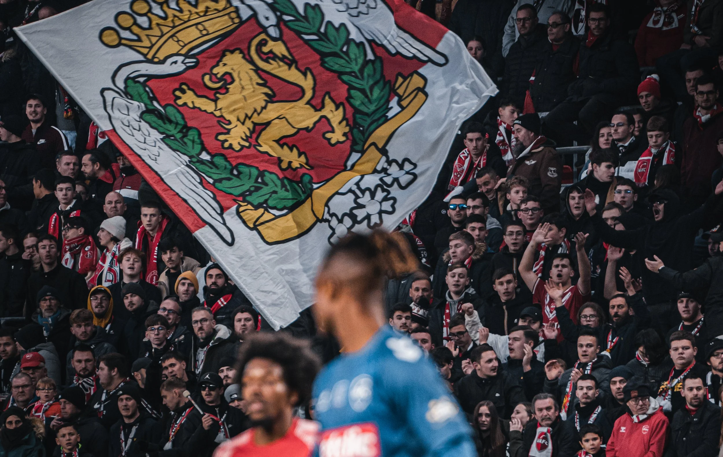 Supporters de football derrière une grande bannière avec un lion doré sur fond rouge, dans un stade