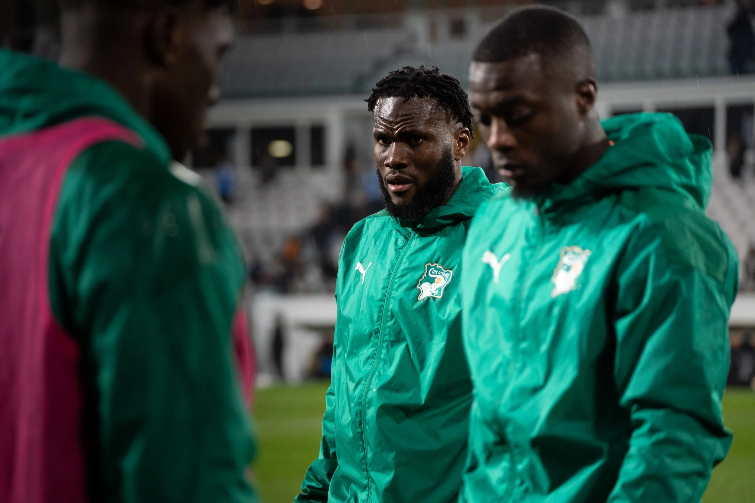 Trois joueurs de football en vestes vertes avec les logos de la Côte d'Ivoire, en discussion sur le terrain.