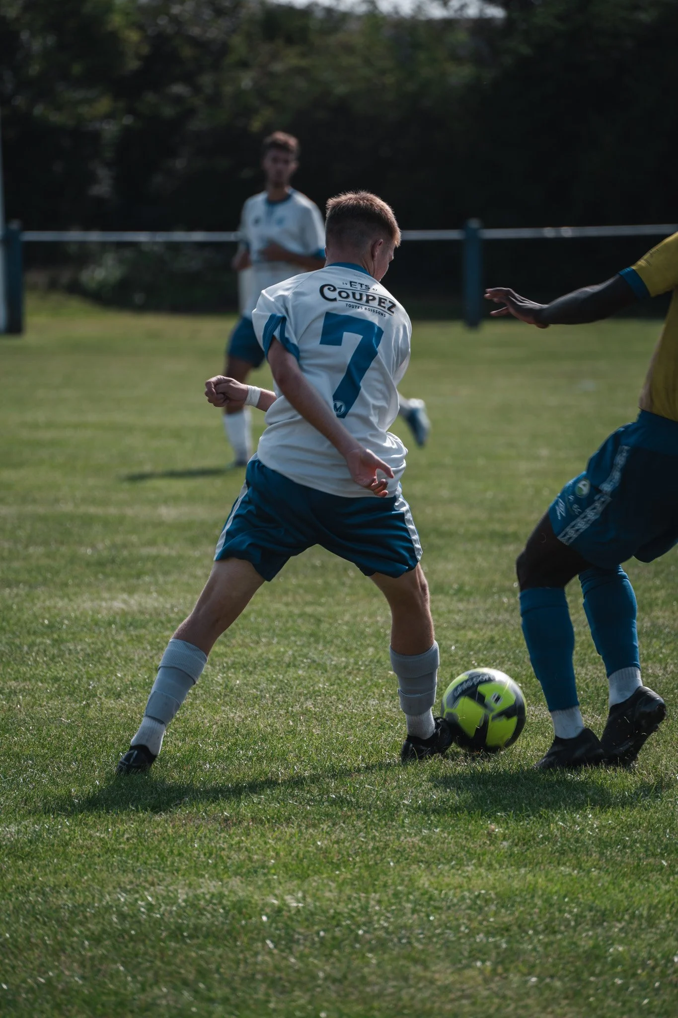 Jeunes joueurs de football en action lors d'un match, l'un d'eux portant un maillot avec le numéro 7 et la phrase "Let’s Coupéz" à l'arrière, en jouant sur un terrain herbeux en extérieur.