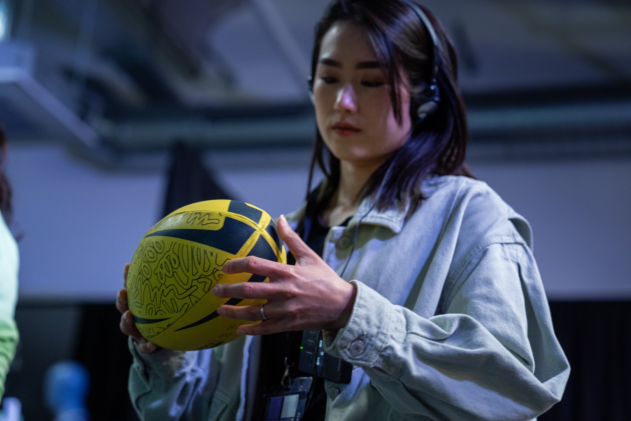 Une femme regarde un ballon de basketball jaune avec des dessins noirs, elle porte un casque ou des écouteurs et une veste en jean. L'arrière-plan est sombre et flou.