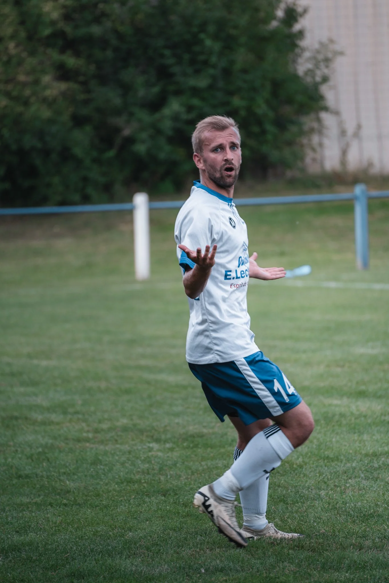 Un joueur de football homme en action sur le terrain, porte un maillot blanc avec le numéro 14, avec une expression confuse ou interrogative.