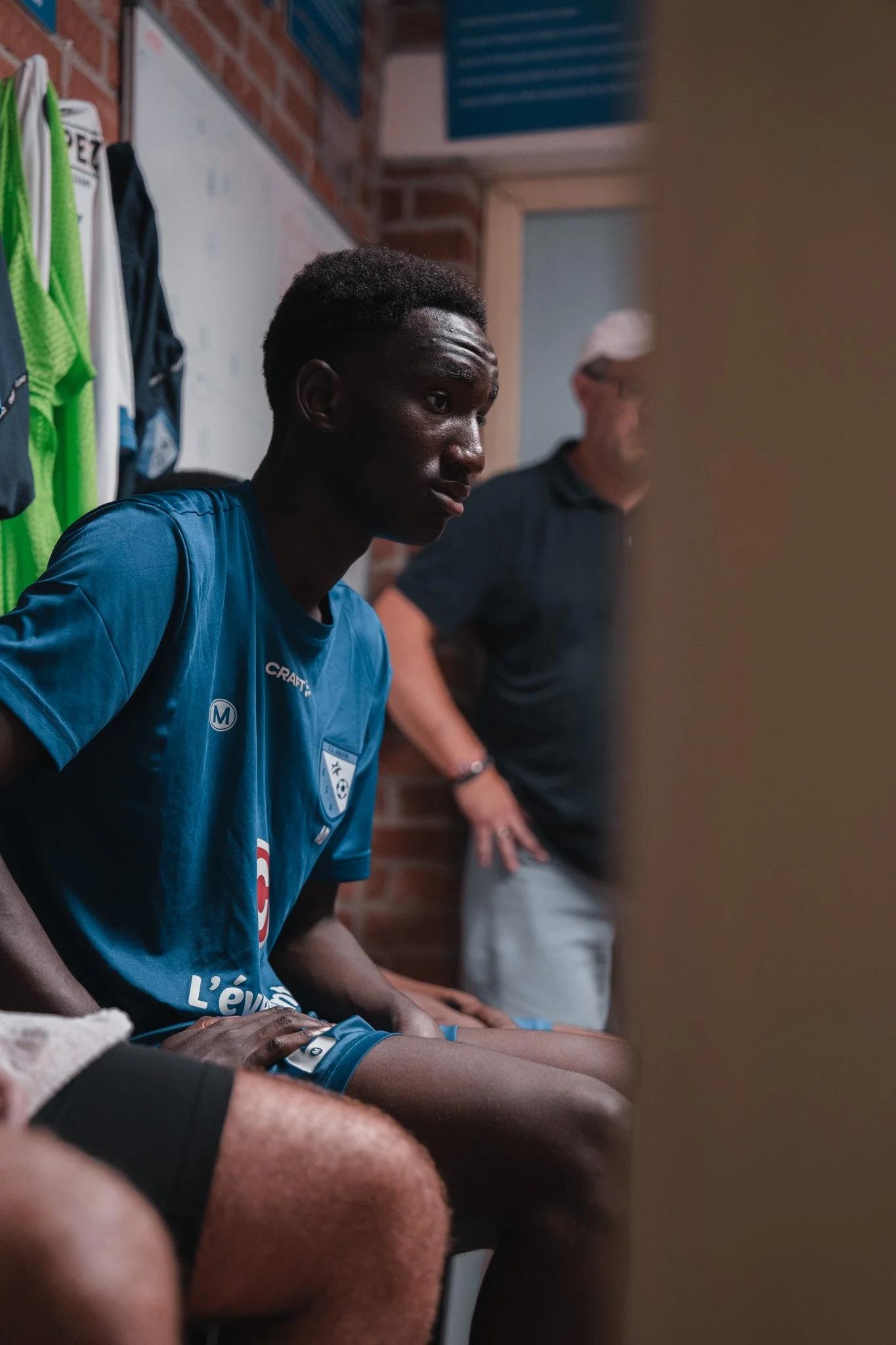 Jeune homme africain en maillot de sport bleu assis dans un vestiaire, avec un adulte en arrière-plan.