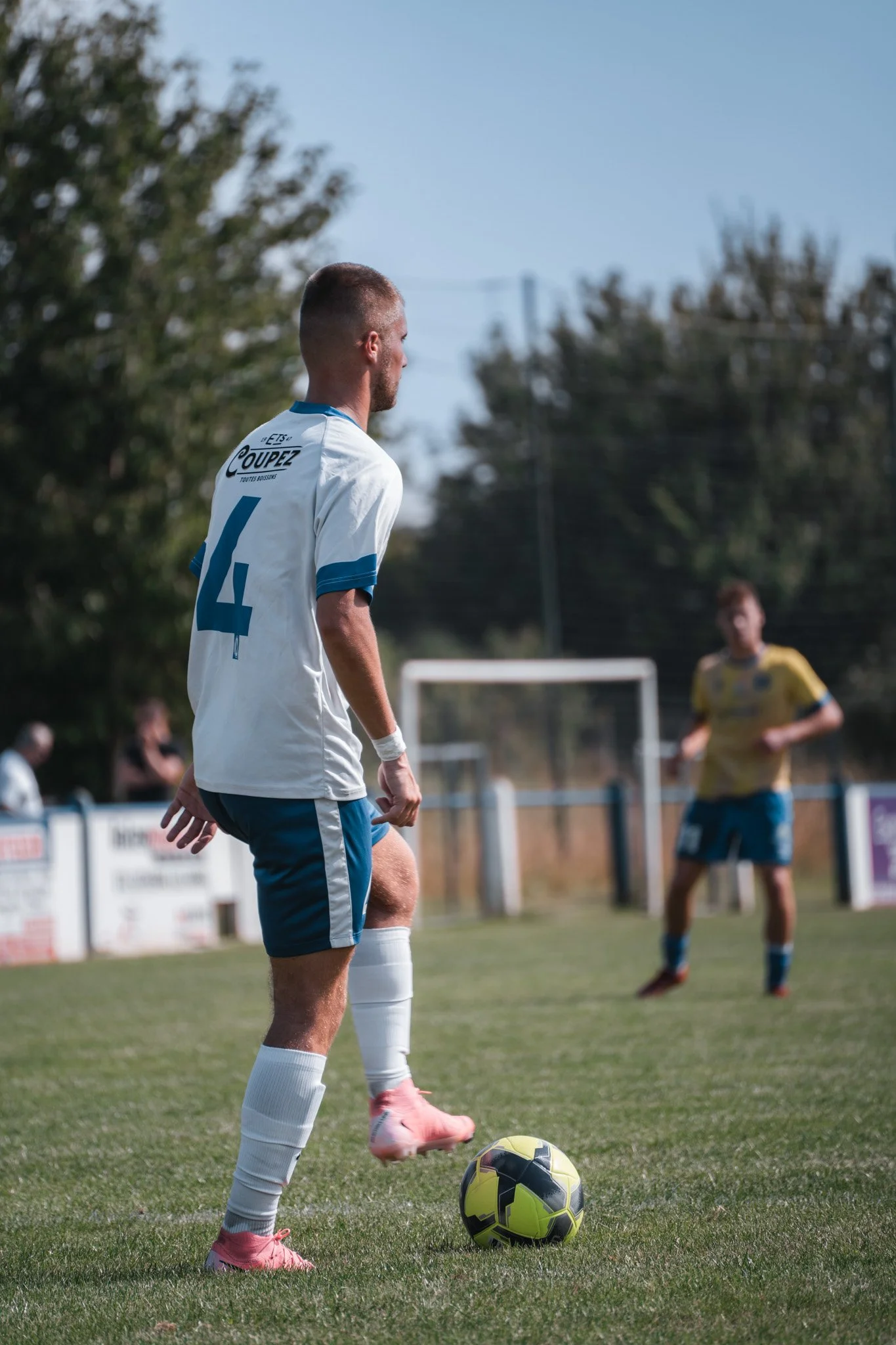 Un joueur de football portant un maillot blanc avec le numéro 4 se prépare à jouer avec un ballon jaune et noir sur un terrain de football, avec un autre joueur en arrière-plan.