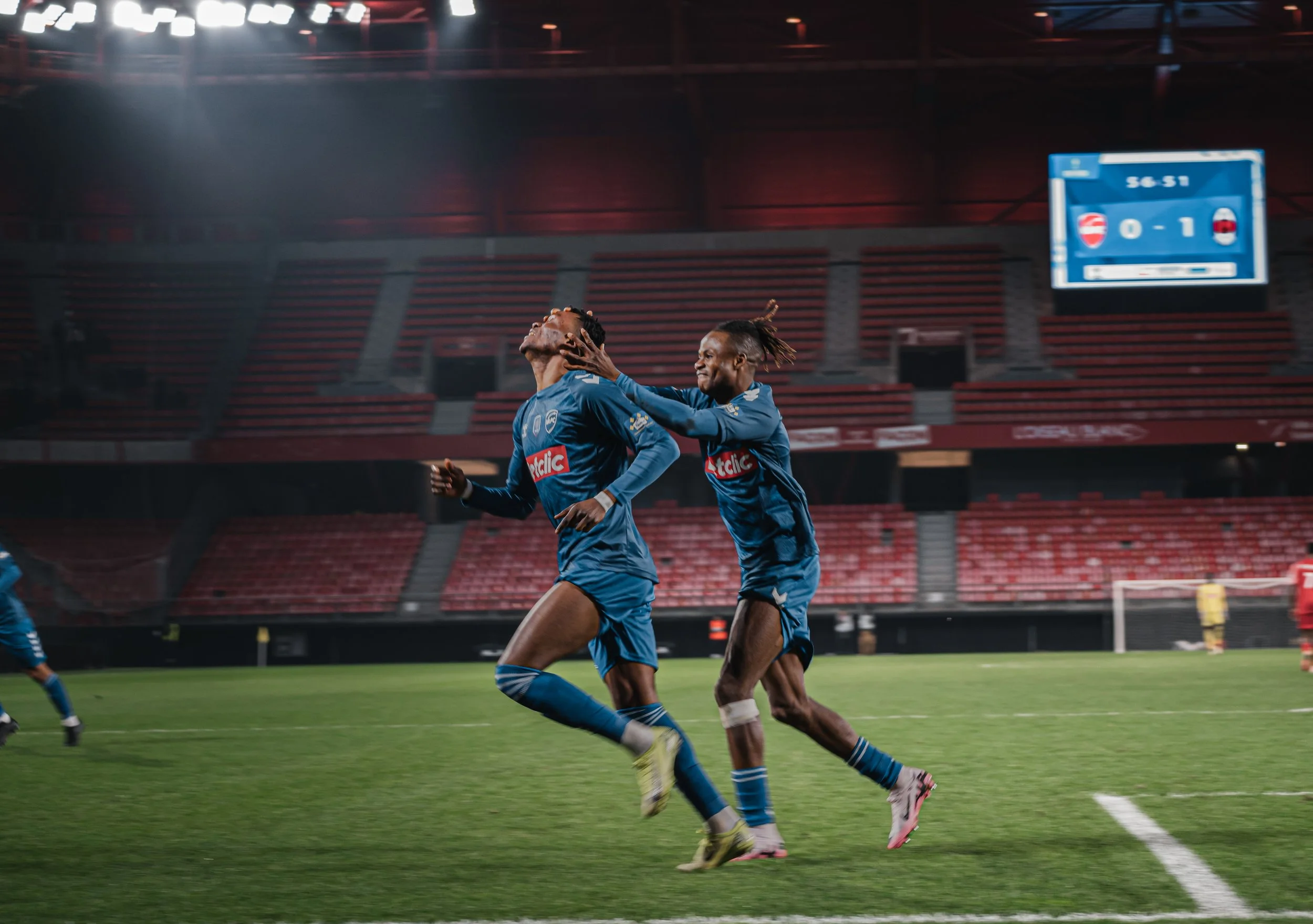 Deux joueurs de football en bleu célèbrent un but sur un terrain de football vide, dans un stade, avec un tableau d'affichage en arrière-plan montrant un score de 0 à 1.