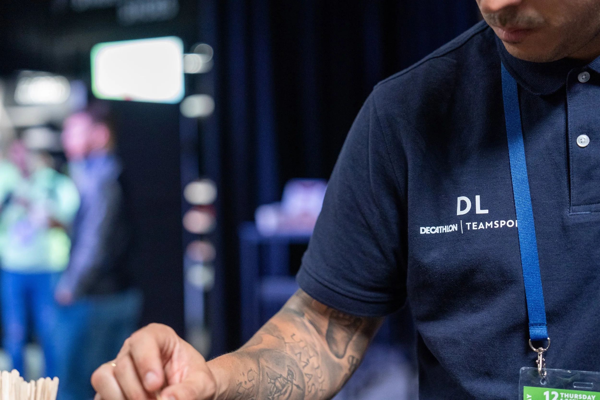 Un homme portant un polo bleu marine avec le logo DECATHLON TEAMSPORT et un badge avec la date 12 jeudi, il a un tatouage sur le bras et porte un lanyard bleu.