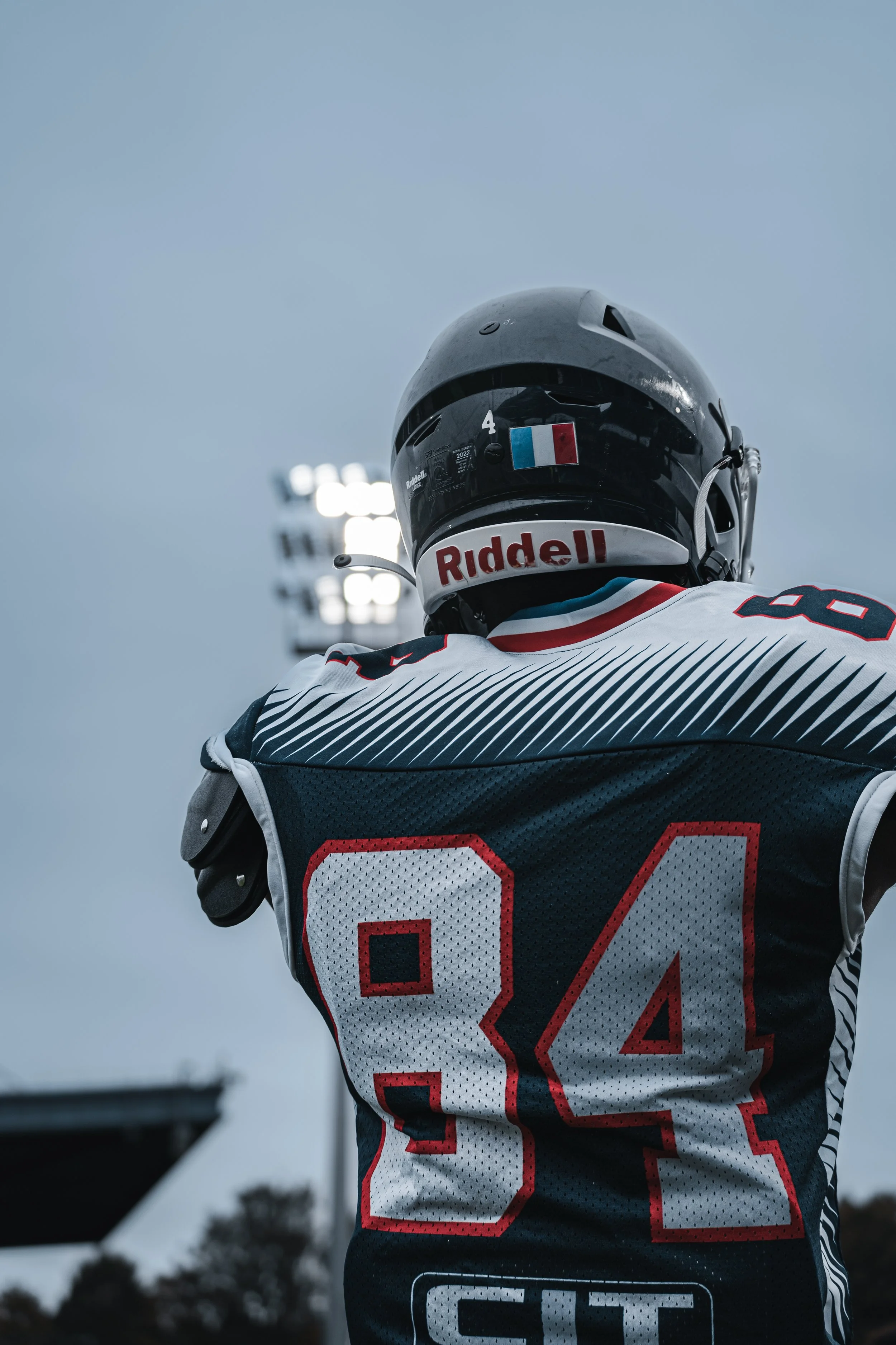 Un joueur de football américain vu de dos portant un casque noir et un maillot avec le numéro 84, sur un fond de ciel nuageux et de terrain