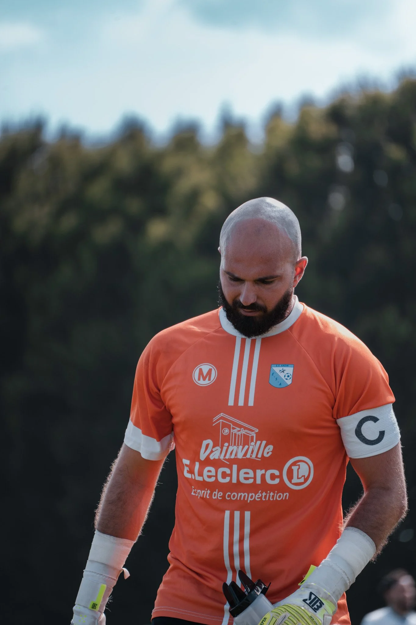 Un joueur de football en maillot orange avec un logo, portant un brassard avec la lettre C, regarde vers le bas sur un terrain.