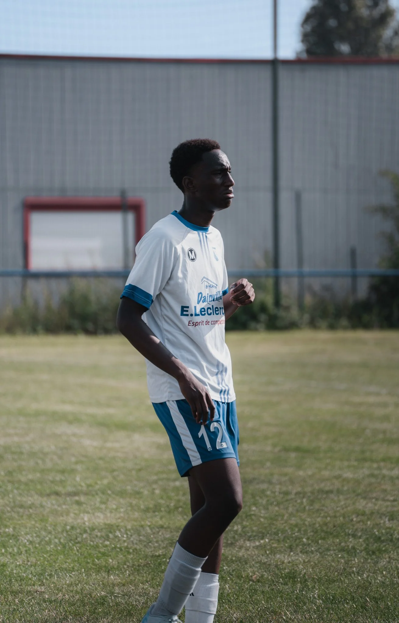 Un joueur de football noir en uniforme blanc et bleu, portant le numéro 12, se trouve sur un terrain de football en plein air.