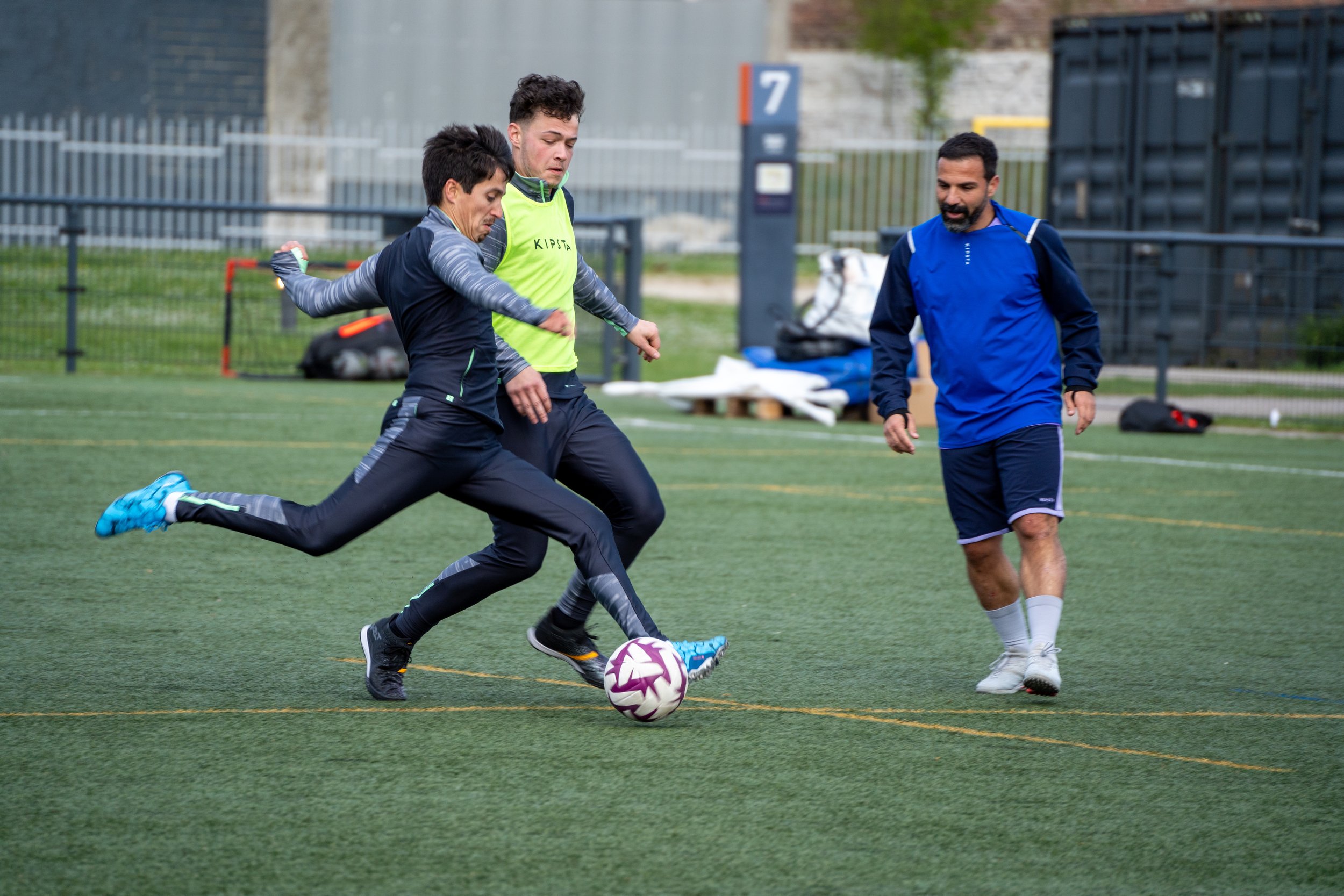 Trois personnes jouent au football sur un terrain en extérieur, deux hommes tentent de prendre le ballon tandis qu'une femme tente de le déloger.