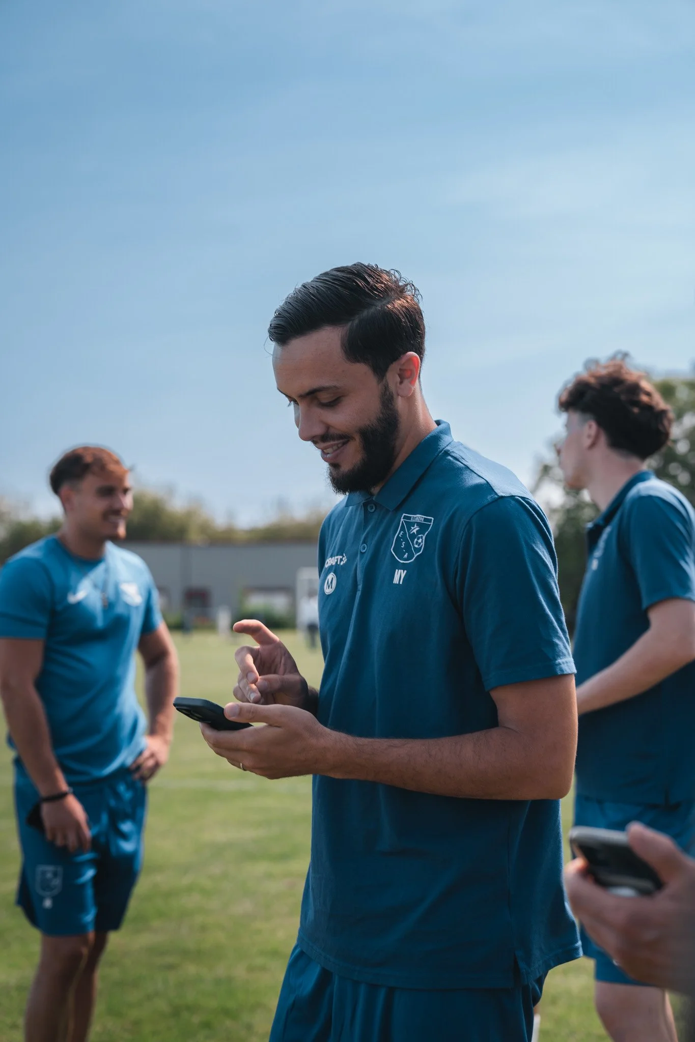 Groupe de jeunes hommes souriants en tenue sportive bleue, échangeant des messages sur leur téléphone portable lors d'une séance d'entraînement ou d'un match de sport en plein air, avec un ciel clair.