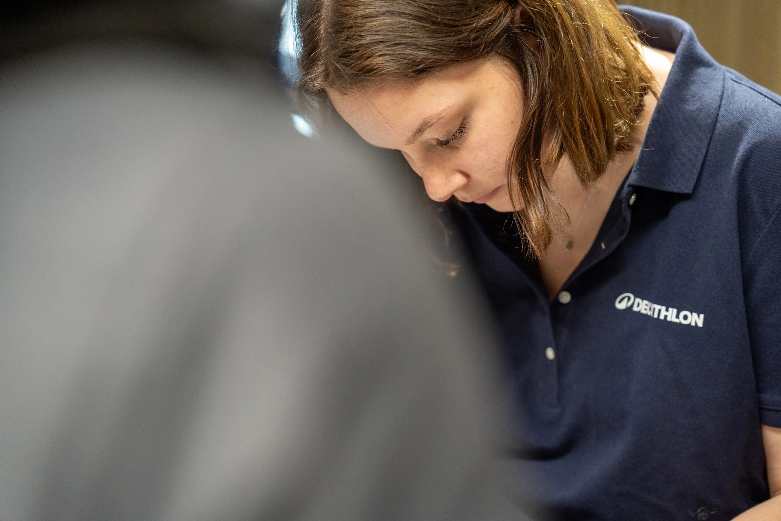 Jeune femme portant un polo bleu avec le logo 'DeThLON', regardant vers le bas, concentrée, dans un environnement intérieur.