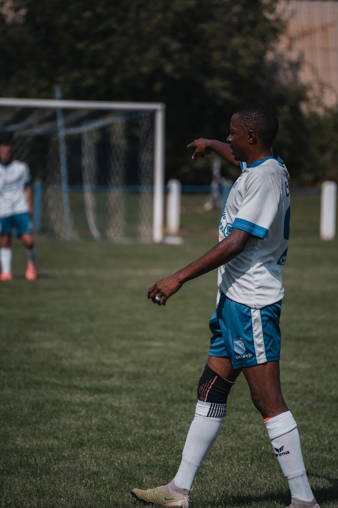 Un joueur de football noir en train de donner un coup de main lors d'un match, portant un maillot blanc et bleu, avec un autre joueur en arrière-plan près du but.