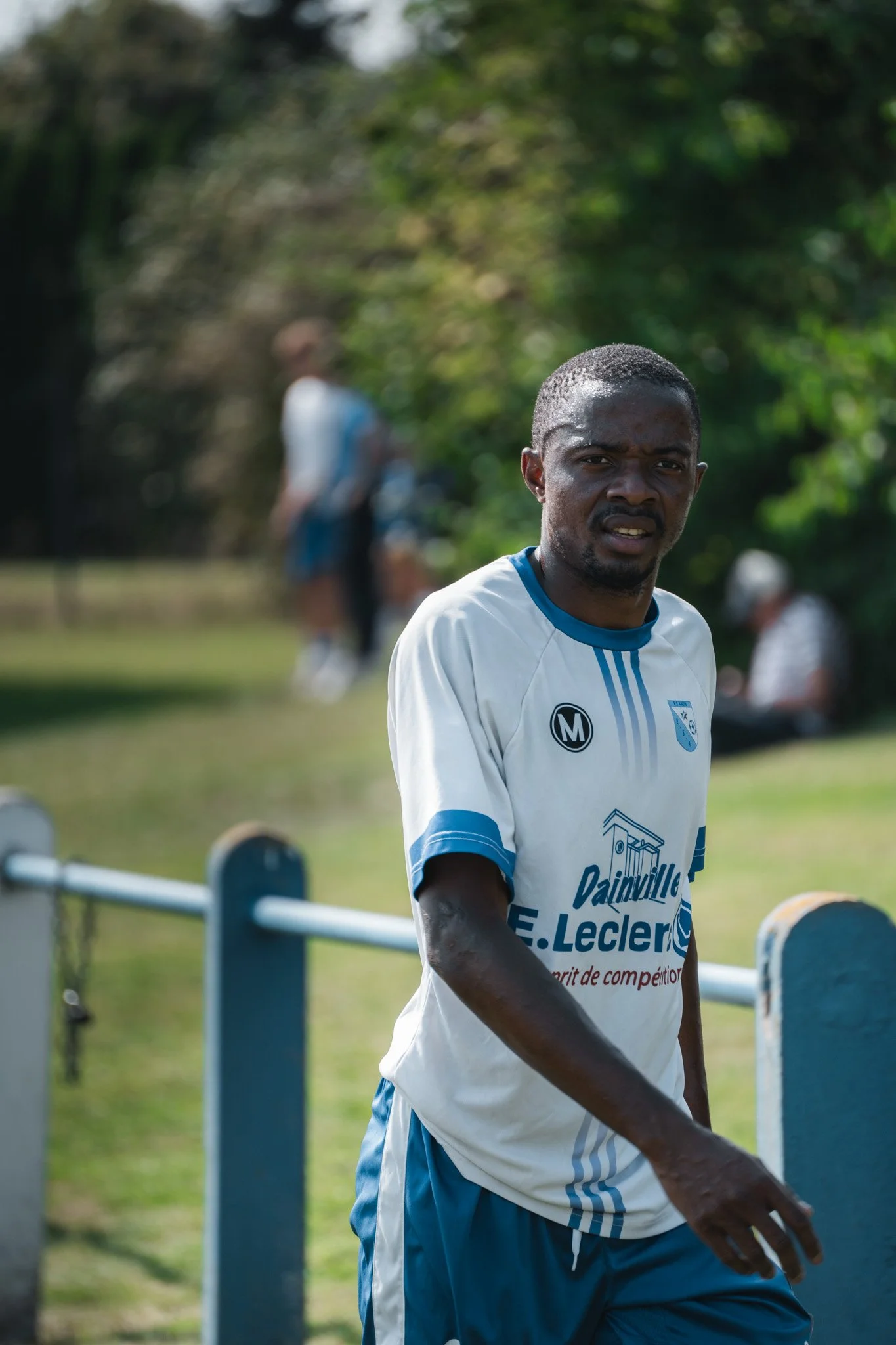 Un homme portant un maillot de sport blanc et bleu, avec des marques et une inscription sur la poitrine, se trouve à l'extérieur. Il a l'air fatigué ou concentré. En arrière-plan, on voit d'autres personnes et un environnement verdoyant et ensoleillé