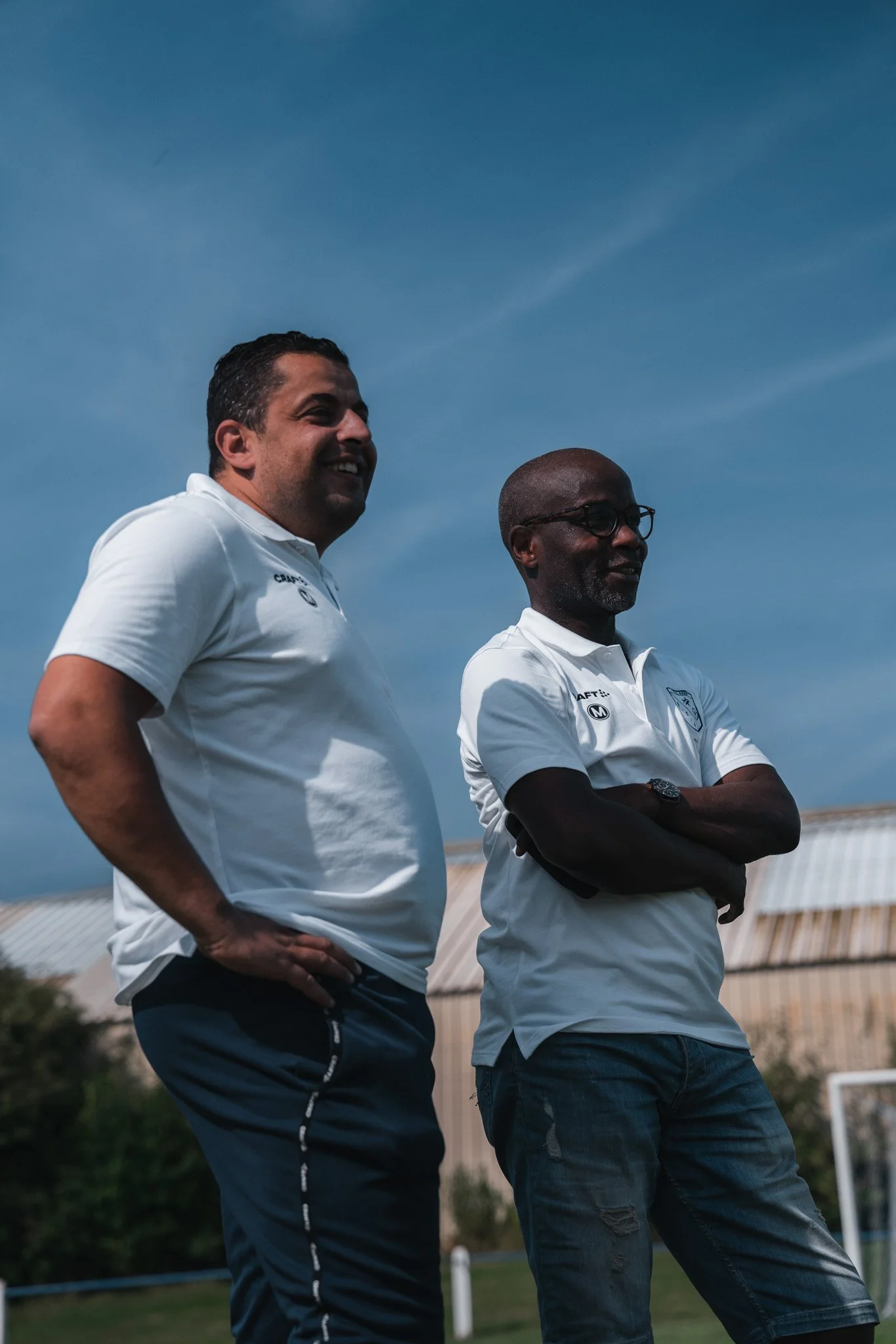 Deux hommes portant des polos blancs sont debout à l'extérieur, avec un bâtiment en arrière-plan, sous un ciel bleu.