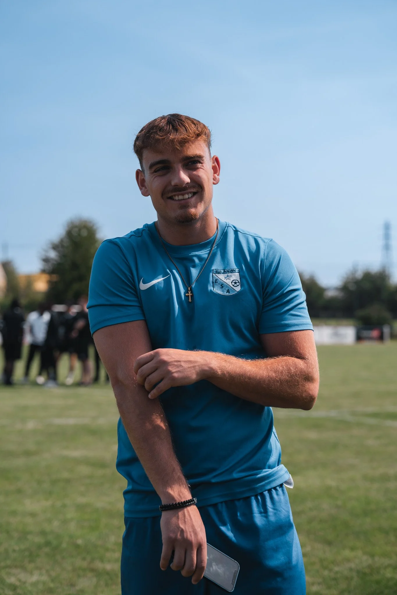 Un jeune homme souriant en tenue de sport bleu, tenant un téléphone portable, sur un terrain herbeux lors d'une activité en plein air sous un ciel dégagé.