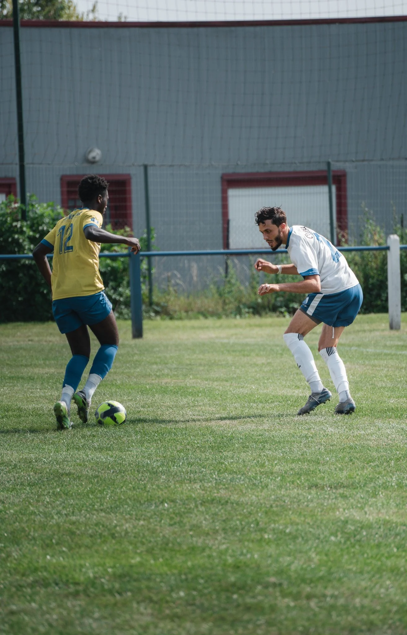 Deux hommes jouent au football sur un terrain herbeux, l'un porte un maillot jaune et les deux portent des shorts et des chaussettes blanches. La scène semble se dérouler lors d'un match ou d'une pratique sportive.