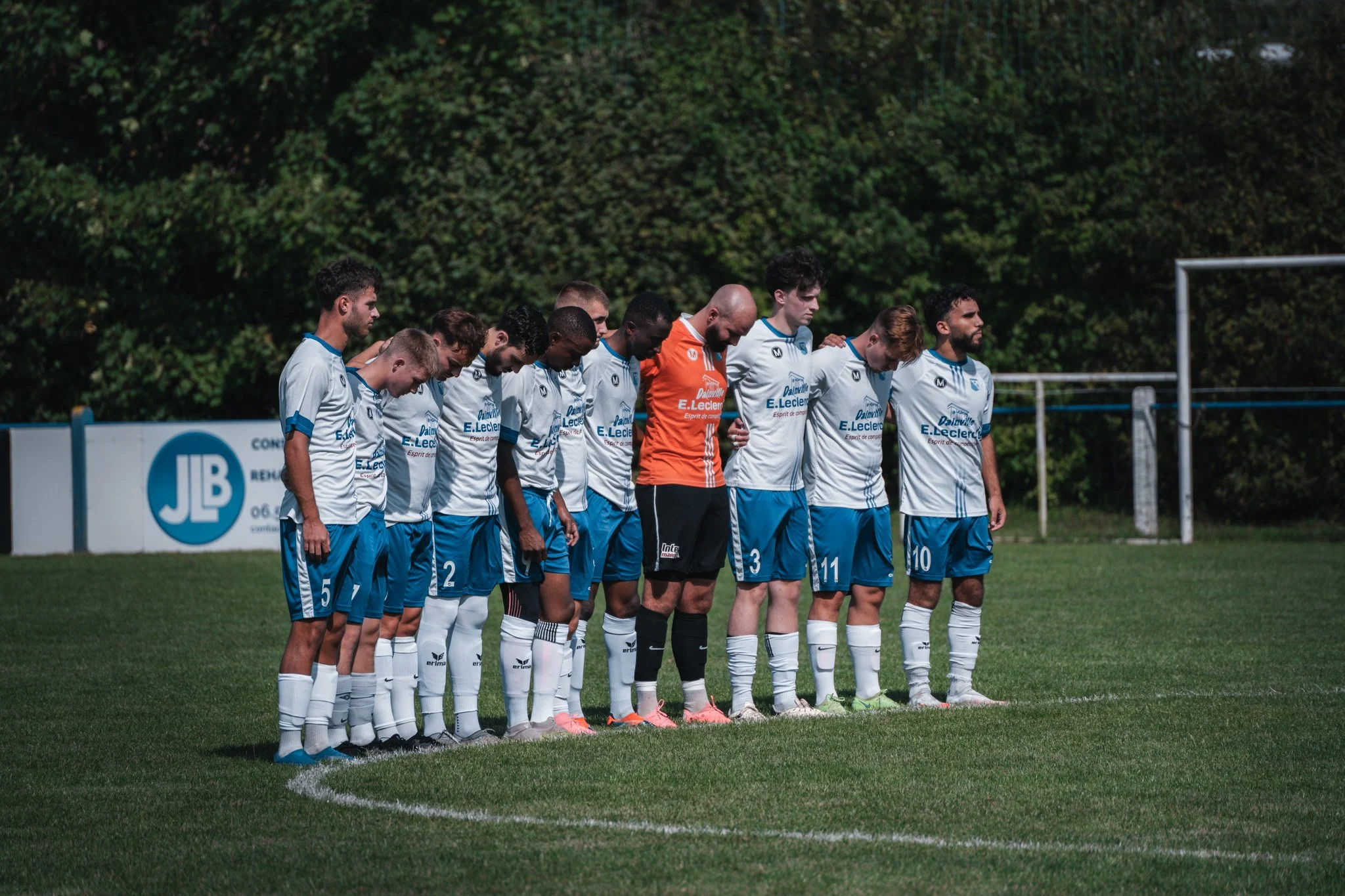 Une équipe de football en ligne avant un match, tous vêtus d'un uniforme blanc et bleu, certains en train de prier ou de se concentrer, sur un terrain vert avec un arrière-plan de feuillage.
