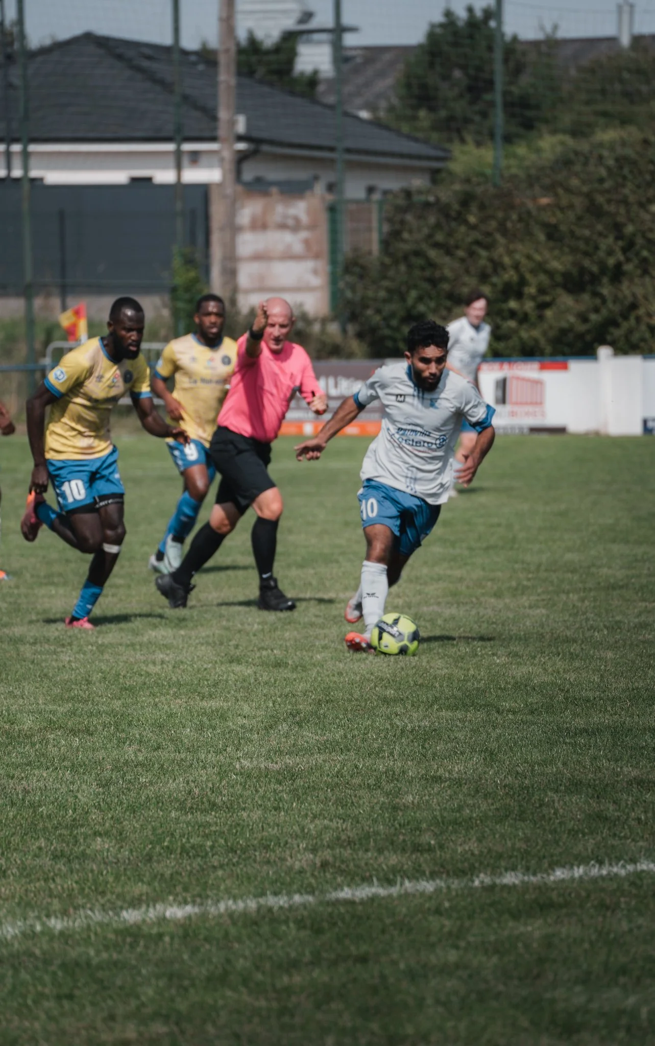 Joueur de football en blanc en train de dribbler avec le ballon sur un terrain. Trois autres joueurs et un arbitre sont derrière lui. La scène se déroule lors d'un match de football en plein air.