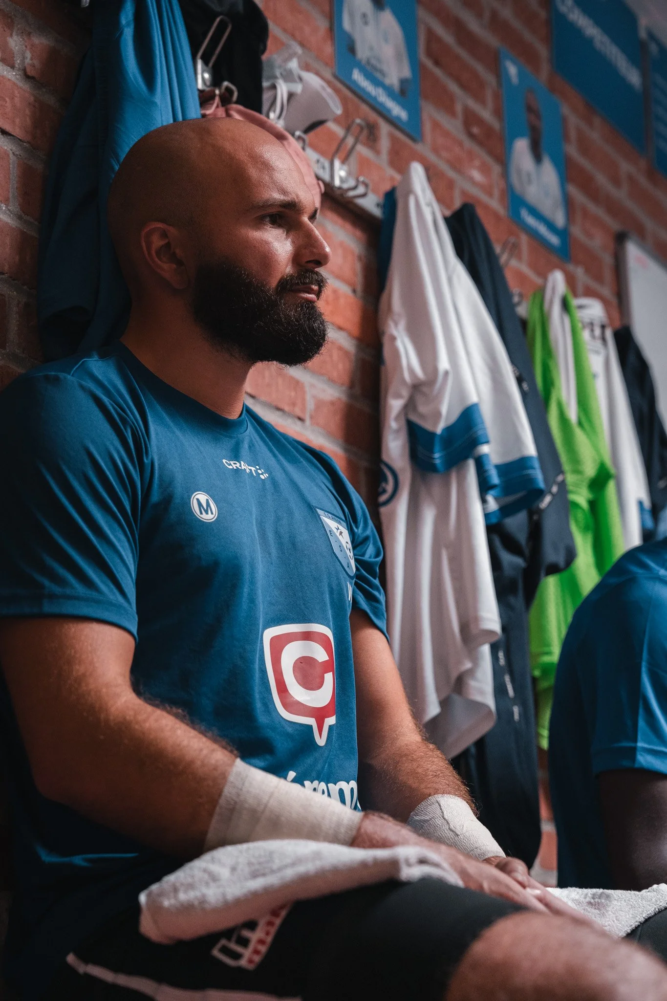 Un joueur de football assis dans un vestiaire, portant un maillot bleu avec un logo et une publicité, avec des vêtements suspendus au mur derrière lui et un mur en briques.