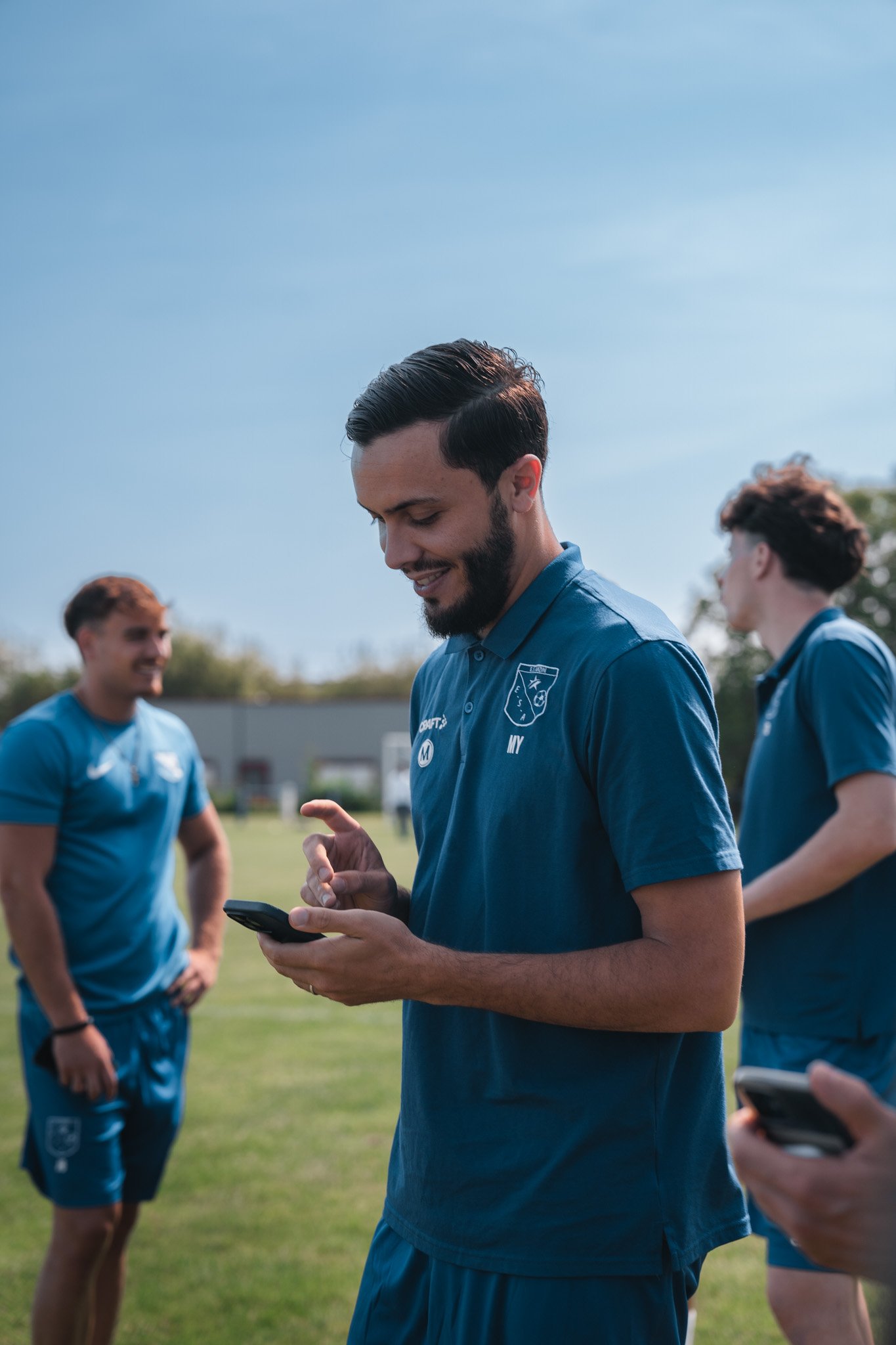 Groupe de jeunes hommes en tenue de sport sur un terrain en herbe en plein air, utilisant des smartphones, sous un ciel bleu clair.