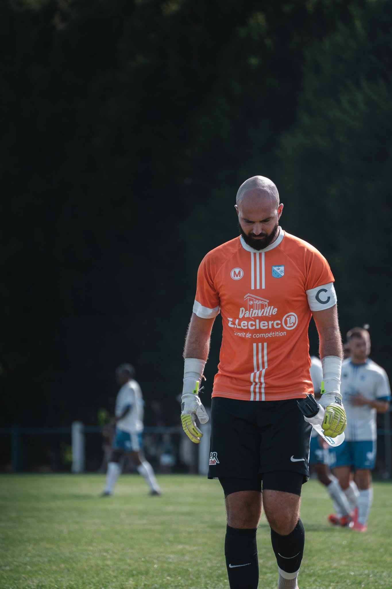 Un joueur de football glabre portant un maillot orange avec un armband 'C', tenant une bouteille d'eau, sur un terrain de sport avec d'autres joueurs en arrière-plan.