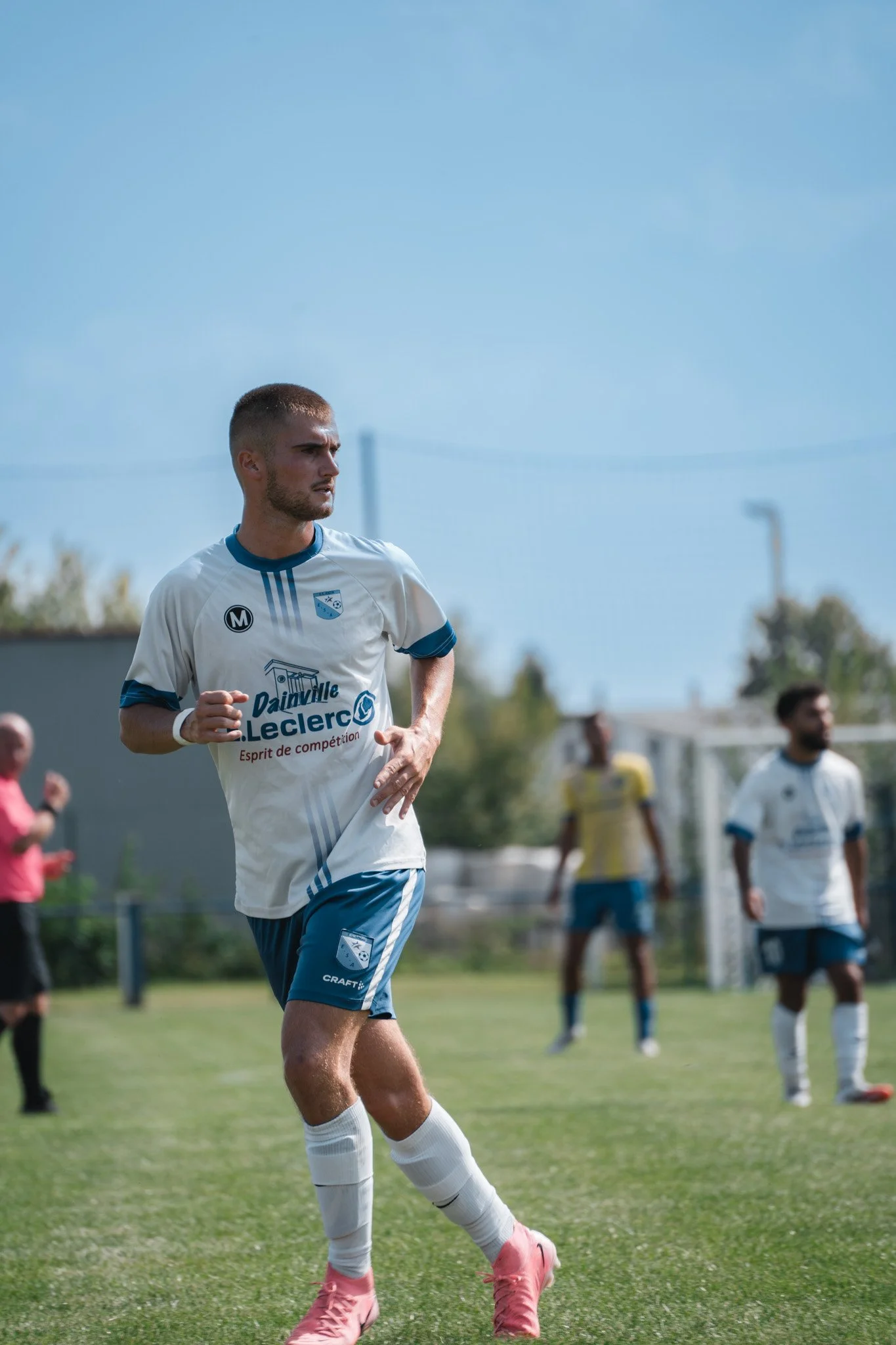 Un joueur de football en train de courir sur le terrain pendant un match, vêtu d'un maillot blanc et bleu avec des sponsors.