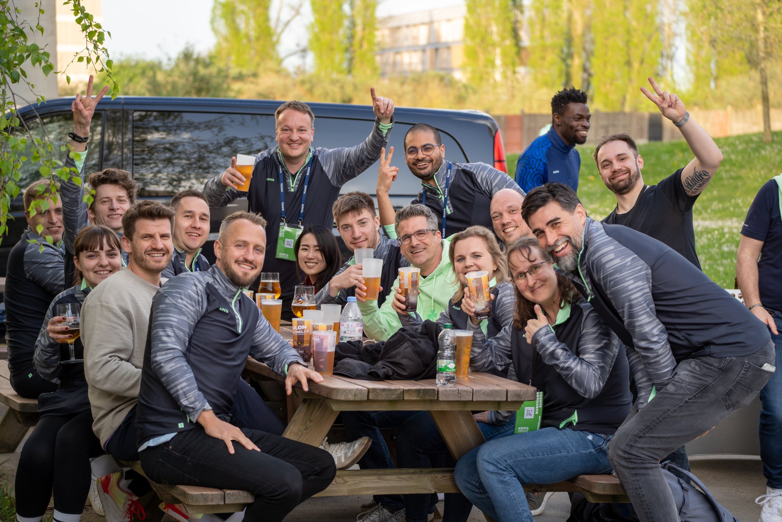 Groupe de personnes souriantes assises et debout autour d'une table en bois, levant des verres de bière lors d'un rassemblement en plein air, avec un véhicule en arrière-plan et des arbres verts.