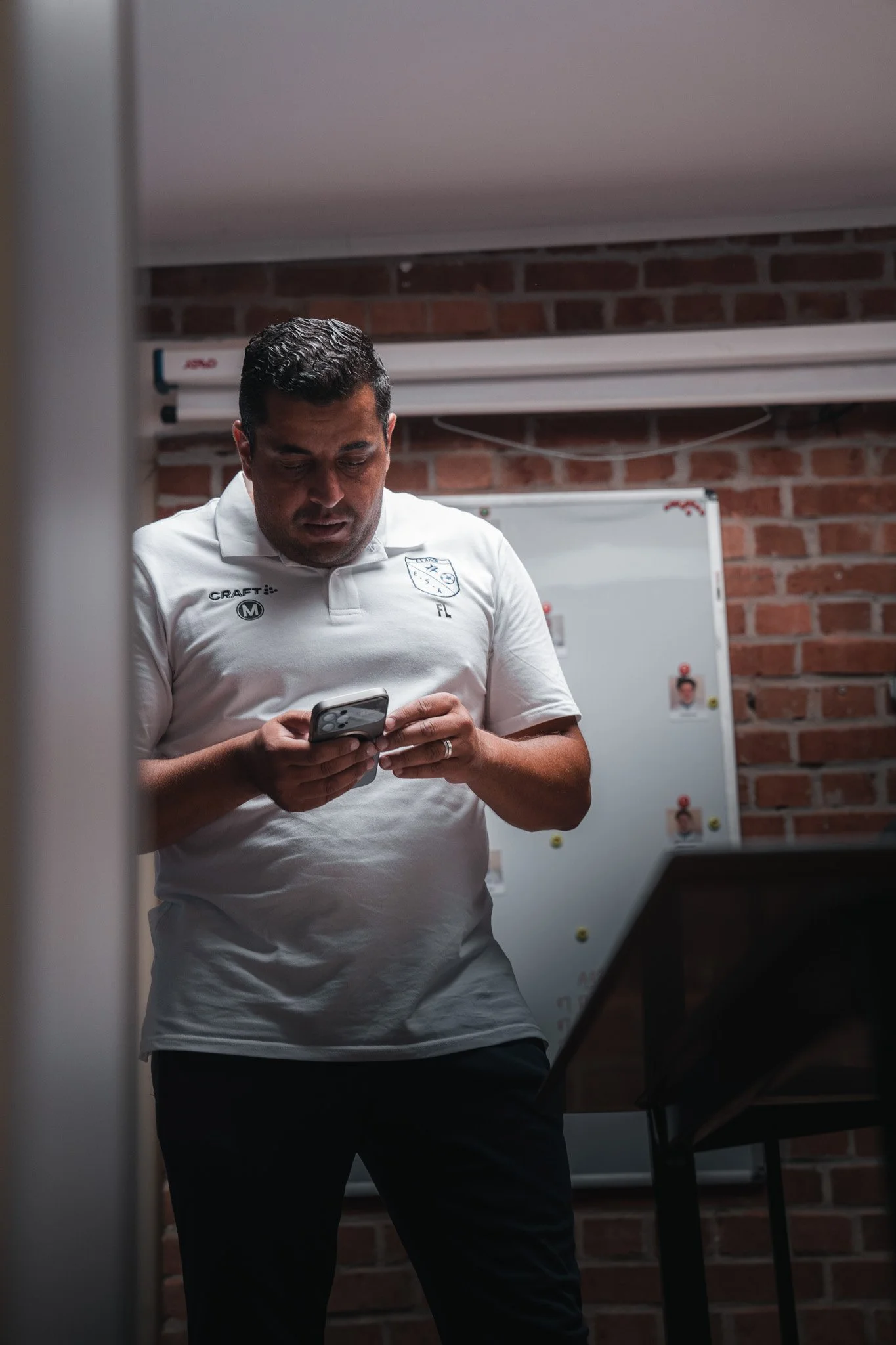 Un homme portant un maillot blanc regarde son téléphone portable dans une pièce avec un mur en briques et un tableau blanc en arrière-plan.