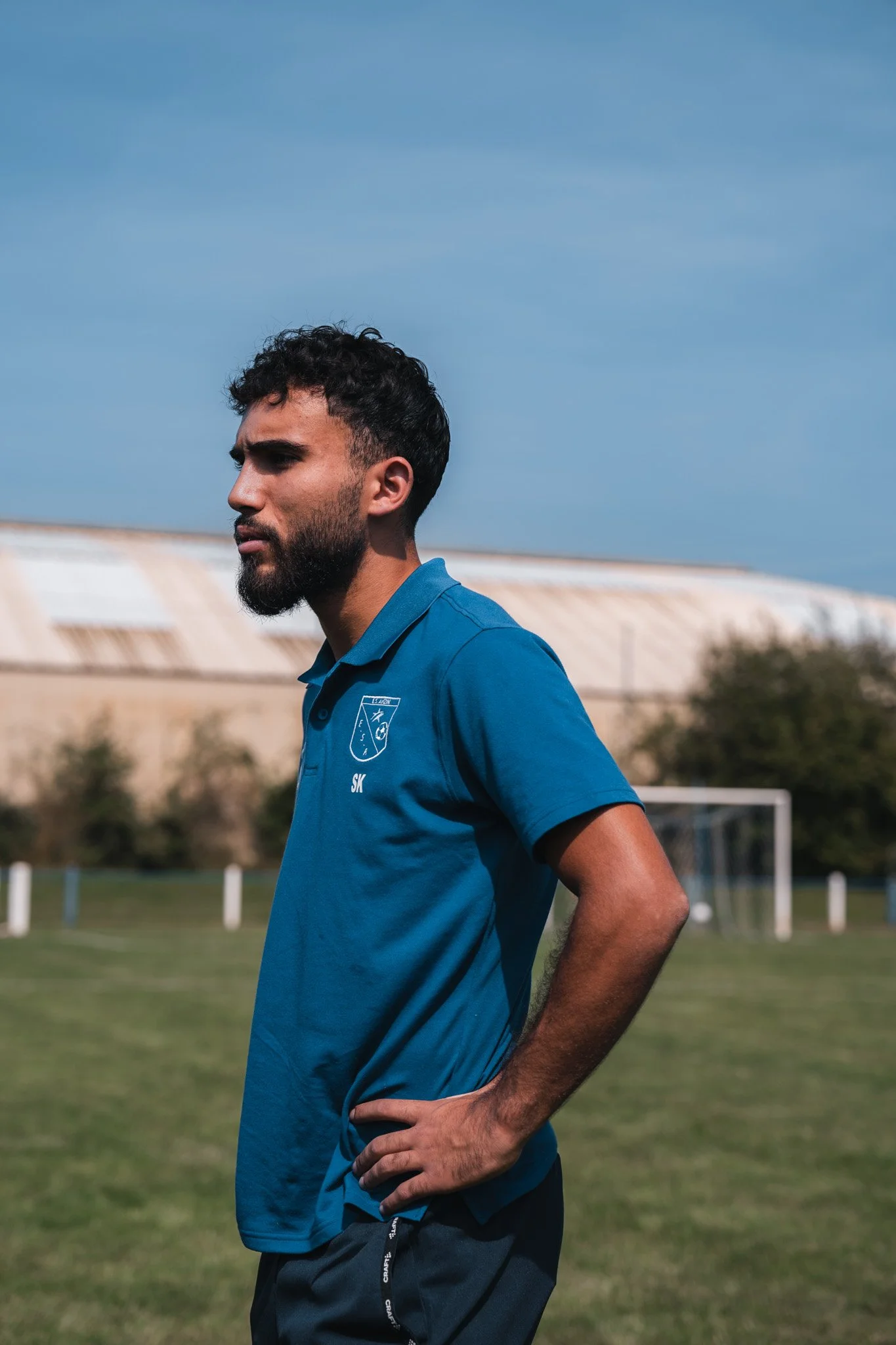 Un homme avec une barbe, tenant sa taille, portant un polo bleu avec un logo d'équipe de football, se tient sur un terrain d'entraînement en plein air avec un but en arrière-plan, sous un ciel bleu.