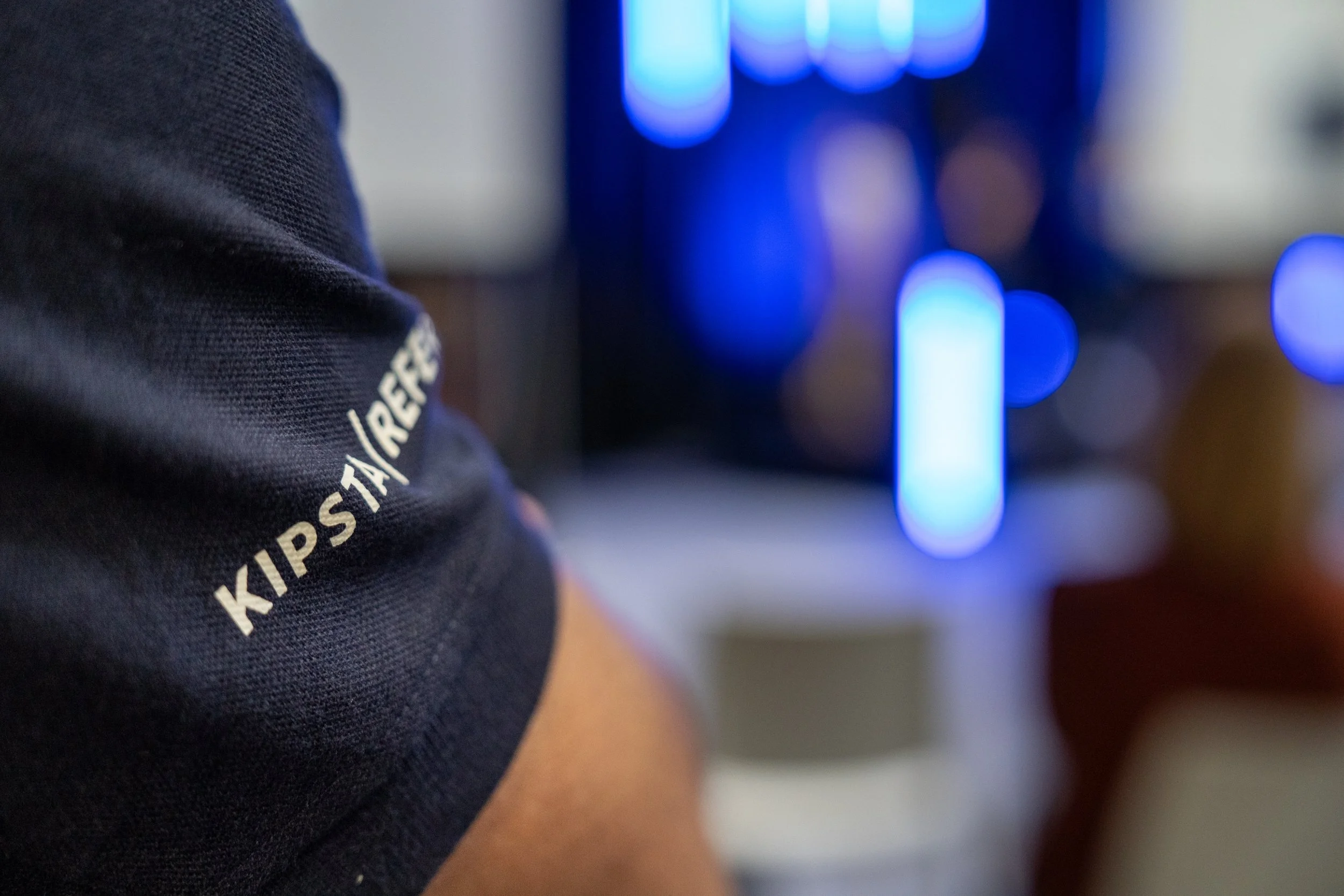 Bras d'une personne portant un T-shirt noir avec l'inscription 'KIPSTA/REUS' et un fond flou avec des lumières bleues.