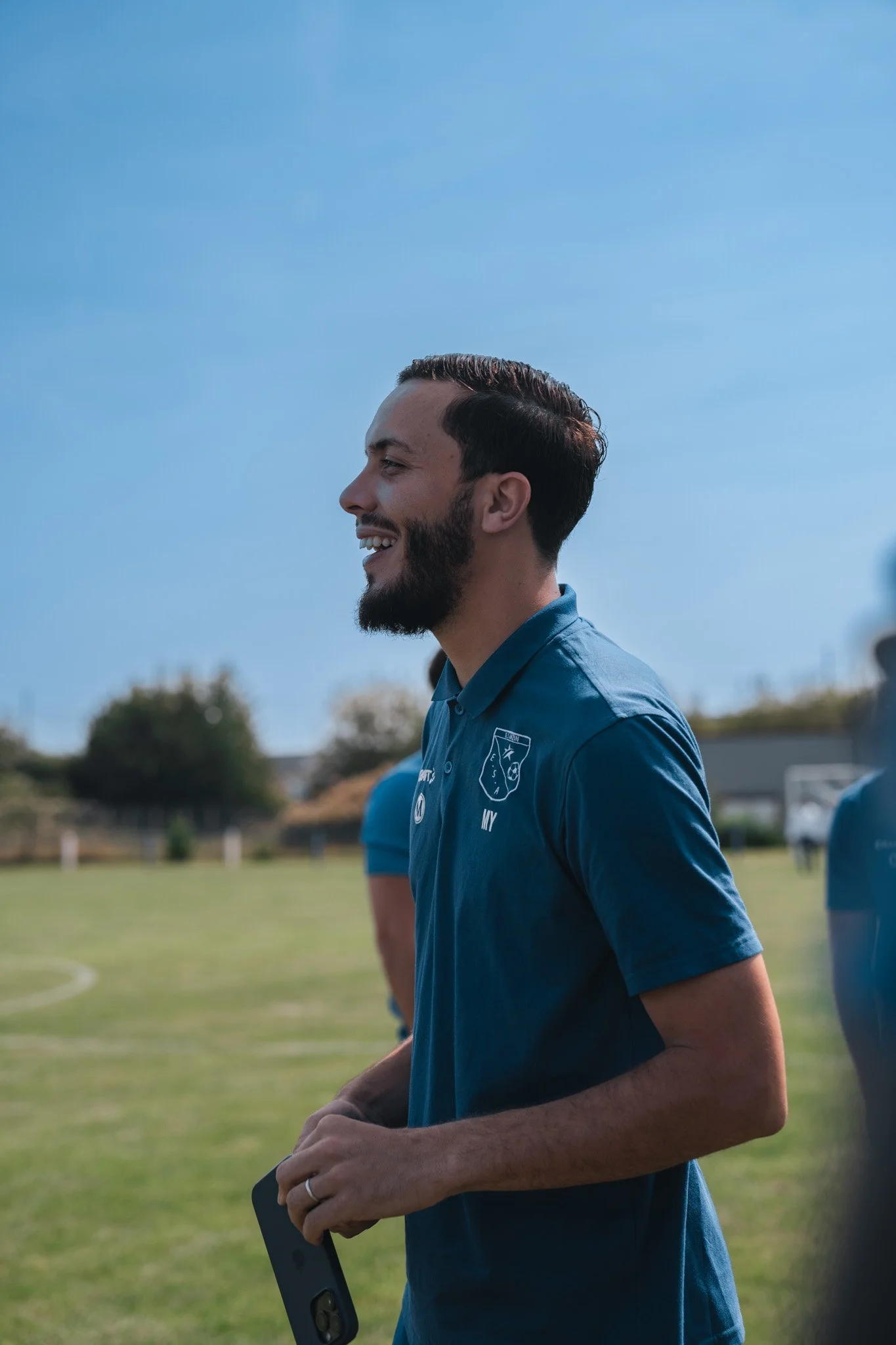 Un homme souriant en tenue de sport sur un terrain de football en plein air, sous un ciel ensoleillé.