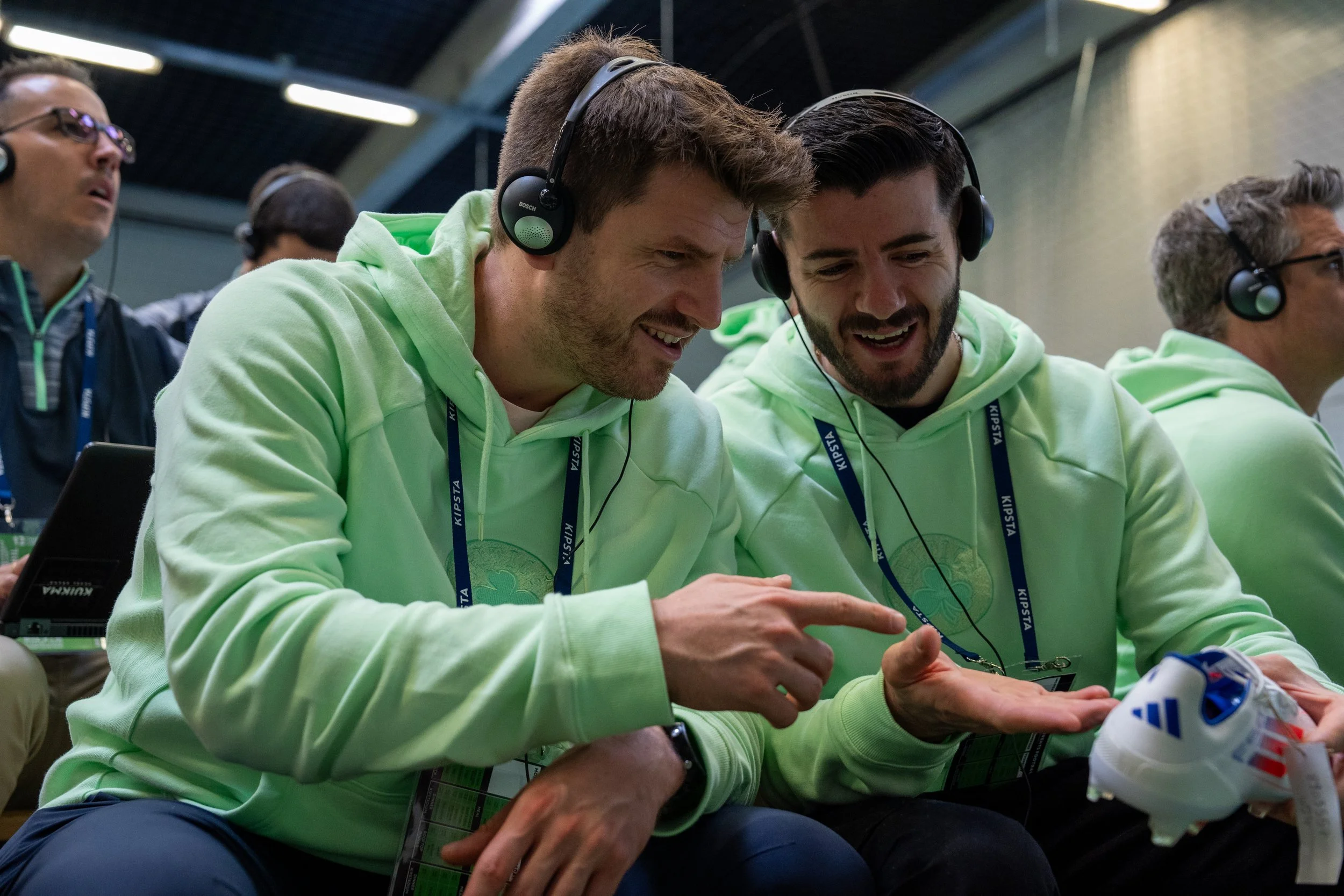 Deux hommes portant des sweat-shirts verts et des écouteurs, souriant et discutant en regardant un objet dans leur main, lors d’un événement sportif ou technologique.