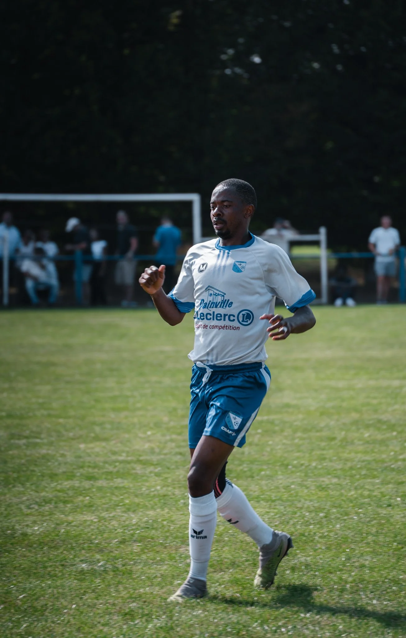 Un joueur de football en tenue blanche et bleue court sur un terrain vert lors d’un match, avec un groupe de spectateurs en arrière-plan.