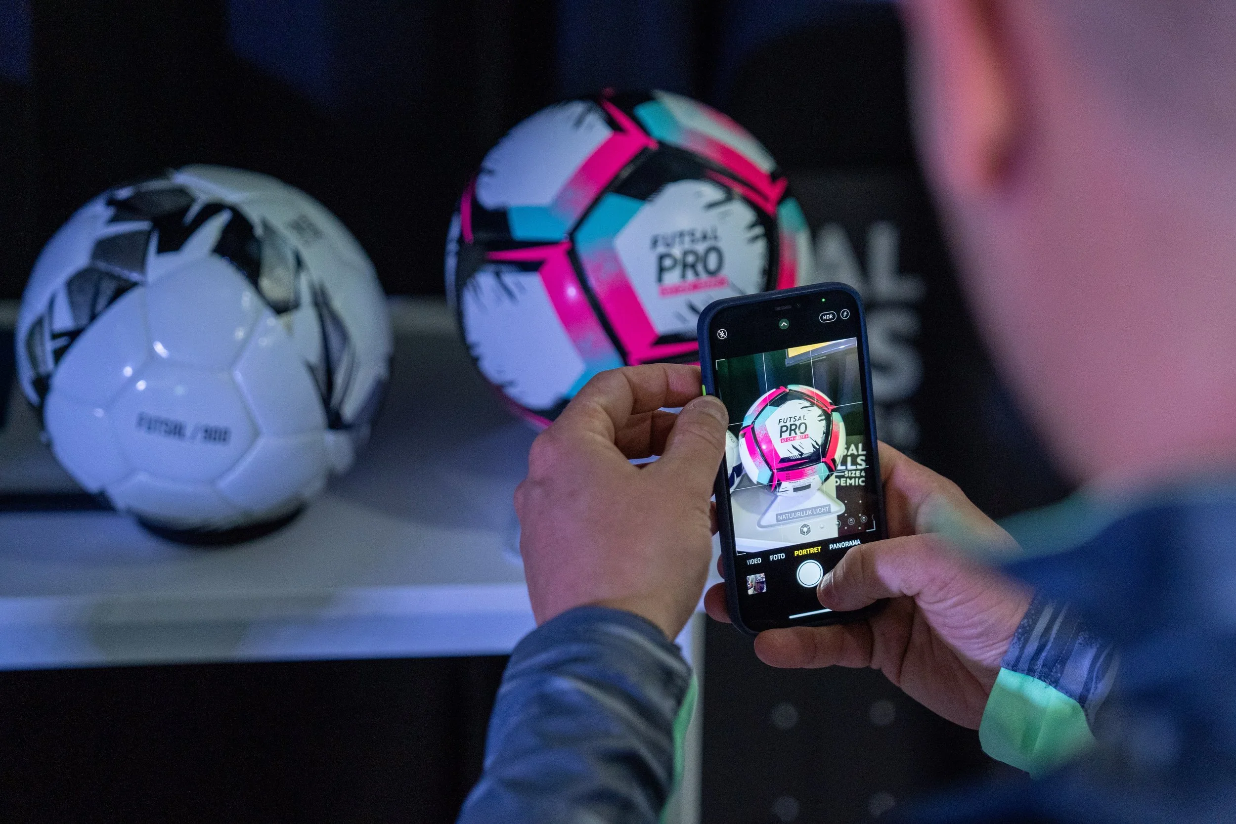 Une personne prend une photo de ballons de football avec son téléphone portable. Les ballons portent le logo 'Futsal Pro' et ont des designs modernes et colorés.