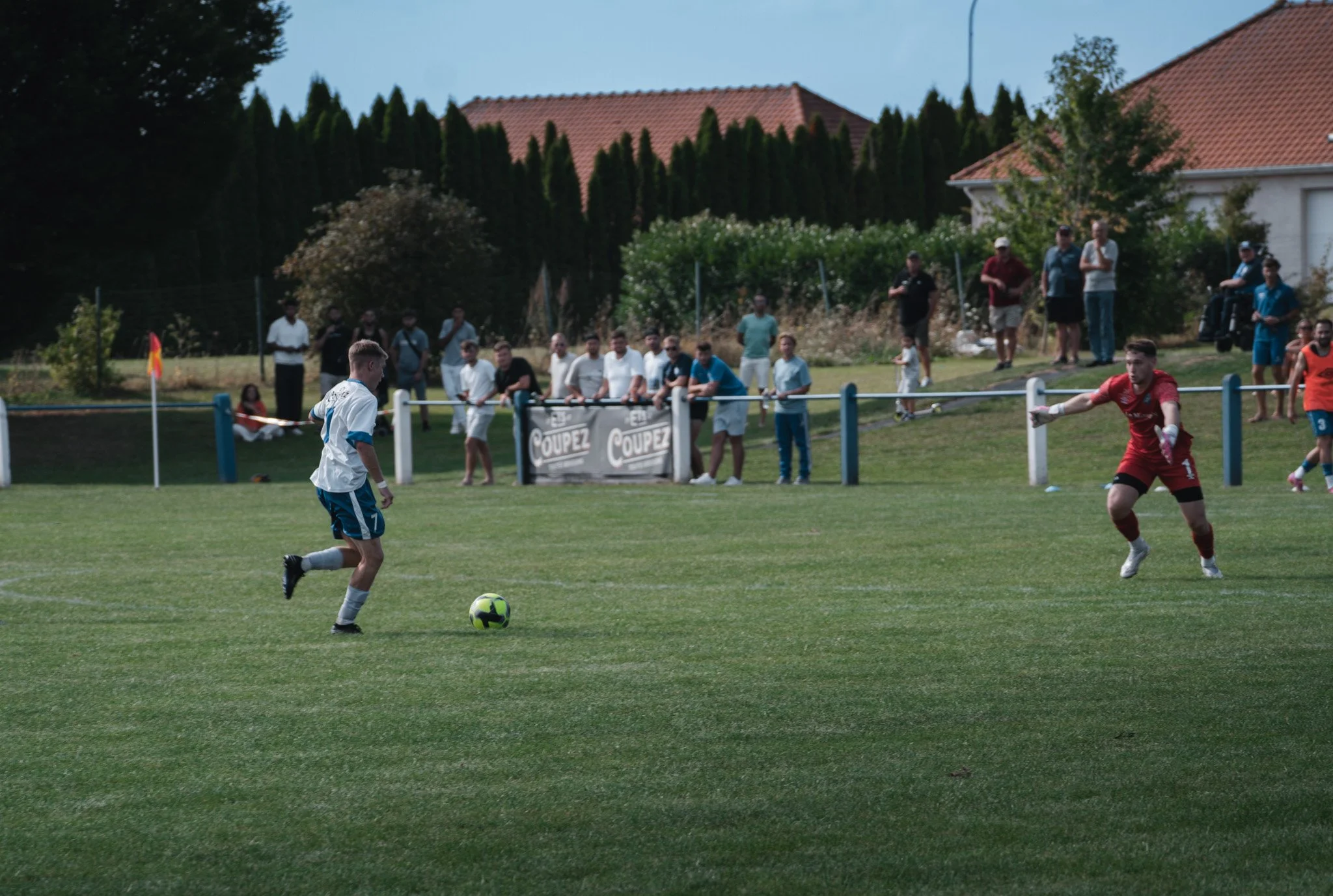 Jeune joueur en blanc et bleu sur un terrain de football, en train de jouer, avec un gardien en rouge face à lui, lors d'un match. Spectateurs et autres joueurs en arrière-plan.