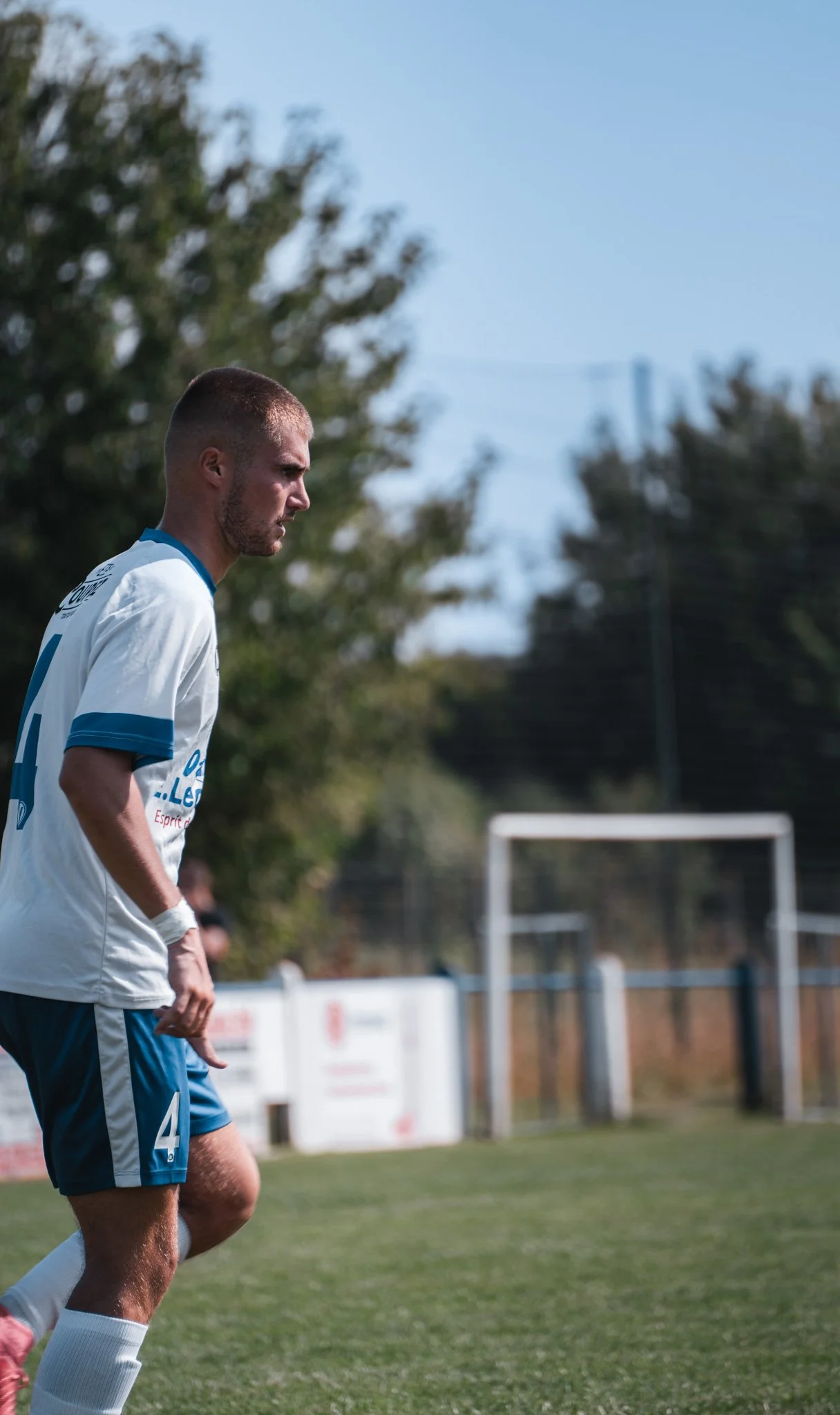 Un joueur de football en uniforme blanc et bleu, l'air pensif, sur un terrain en plein air.