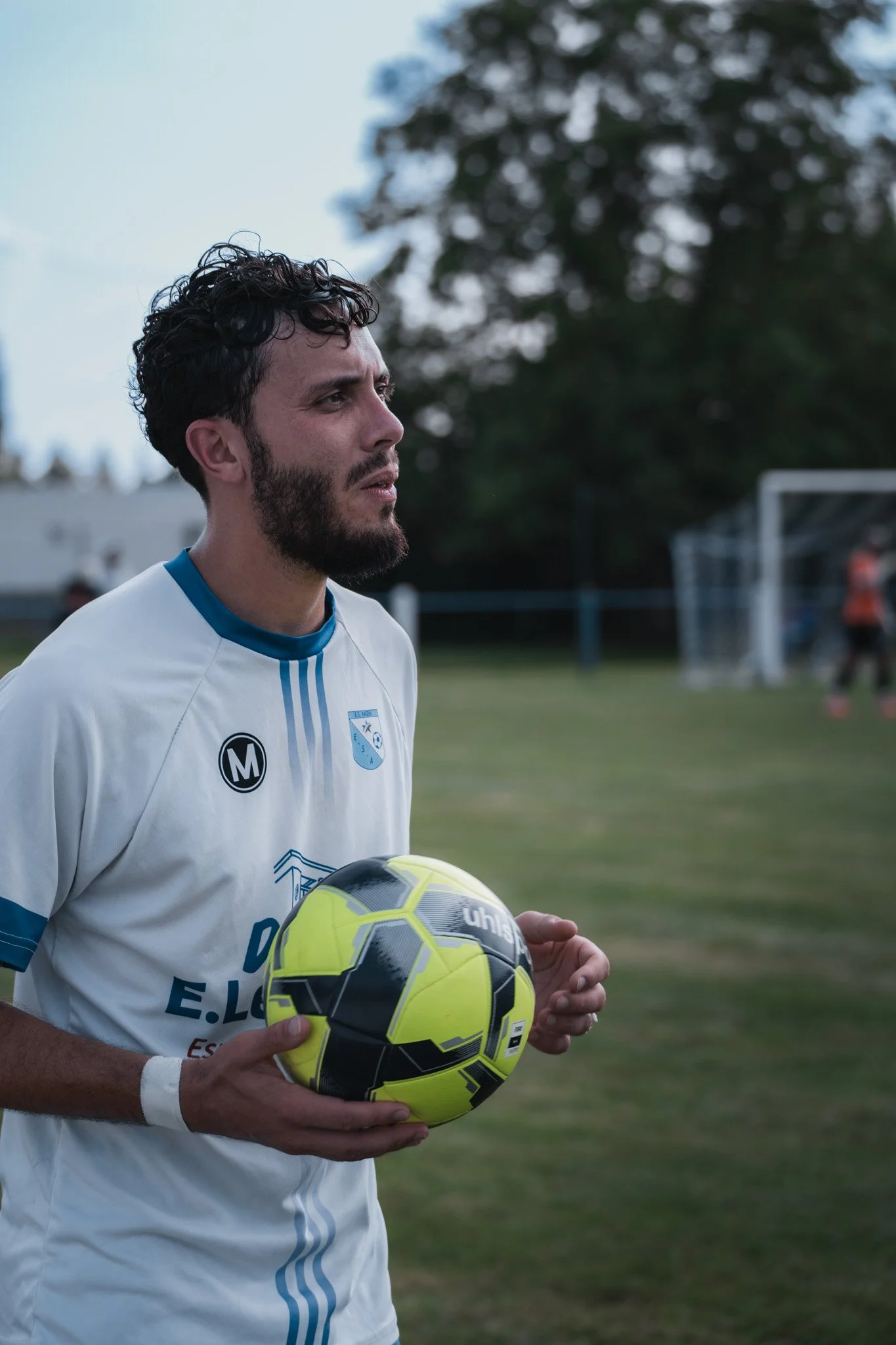 Un homme avec une barbe tient un ballon de football jaune et noir sur un terrain de sport. Il porte un maillot blanc avec des rayures bleues et un logo.
