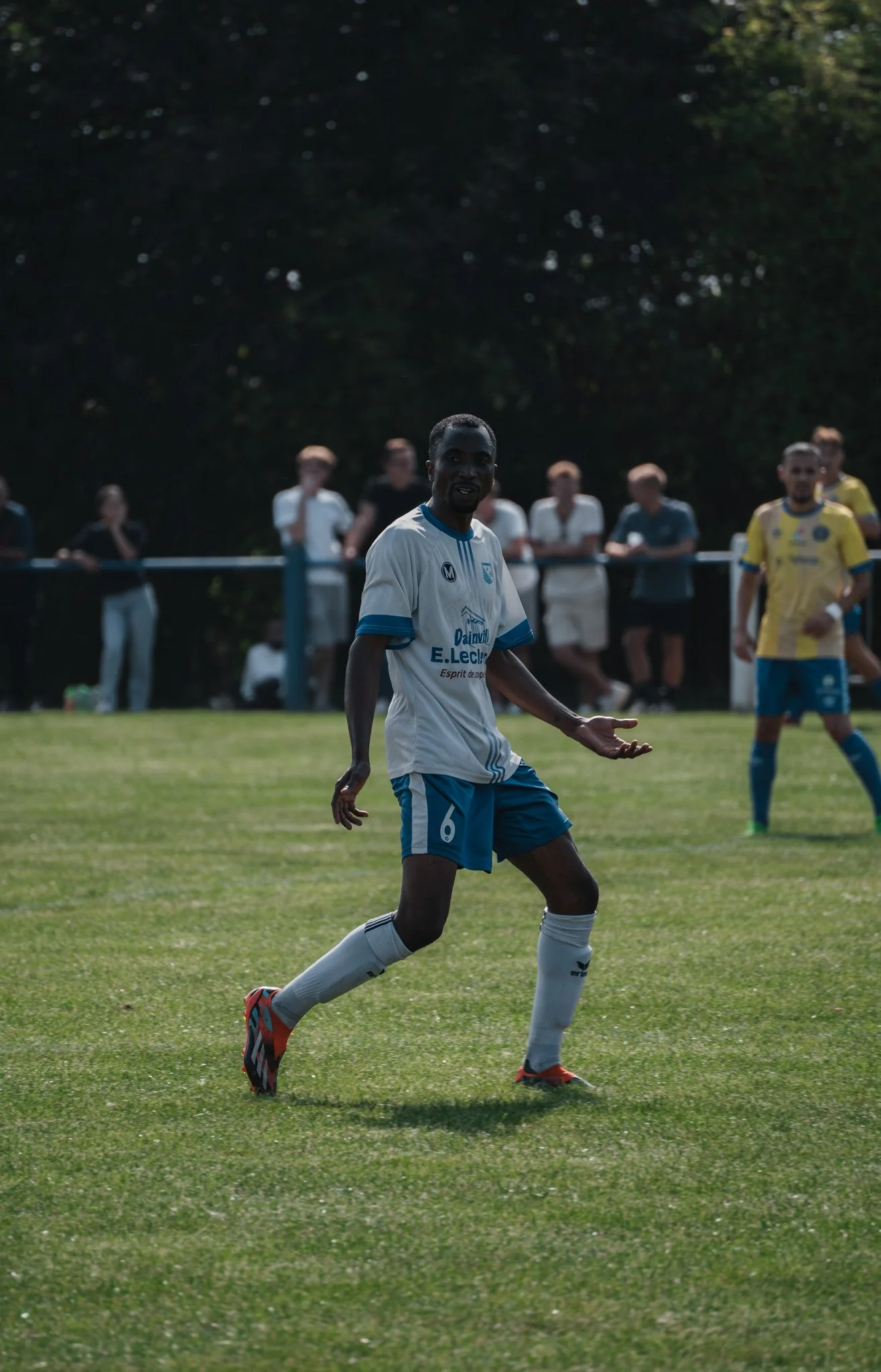 Un joueur de football noir portant un maillot blanc avec des rayures bleues, des shorts bleus, et des chaussettes blanches, en train de jouer sur un terrain de football, avec des spectateurs en arrière-plan.