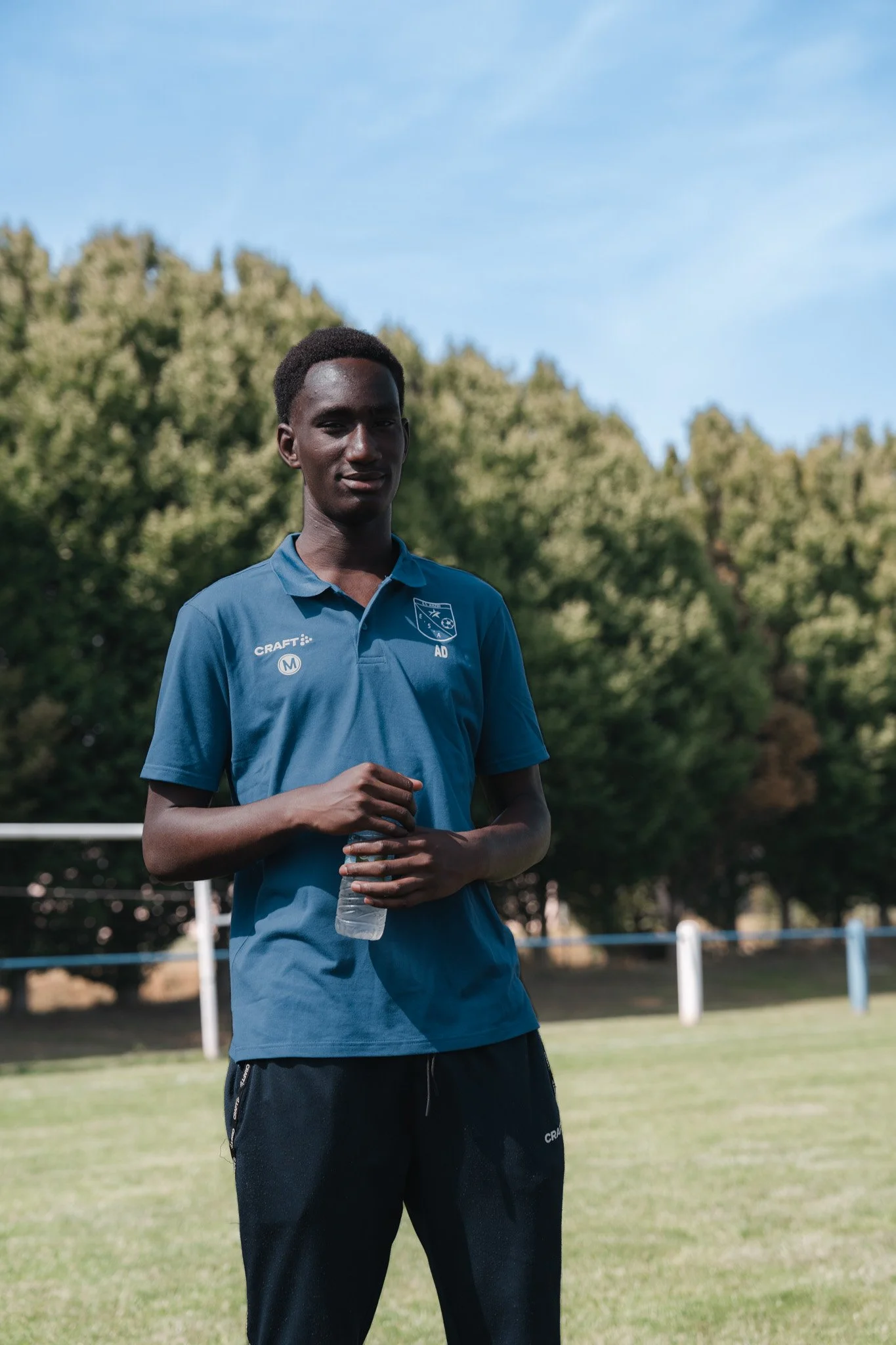 Jeune homme portant un polo bleu avec le logo d'une équipe de sport, debout dans un terrain de sport avec des arbres en arrière-plan, tenant une bouteille d'eau.