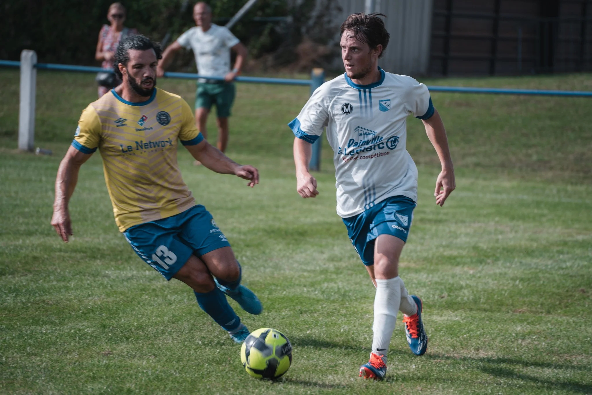 Deux joueurs de football en compétition sur le terrain, l'un en maillot jaune et l'autre en maillot blanc, poursuivant le ballon dans un match amateur.