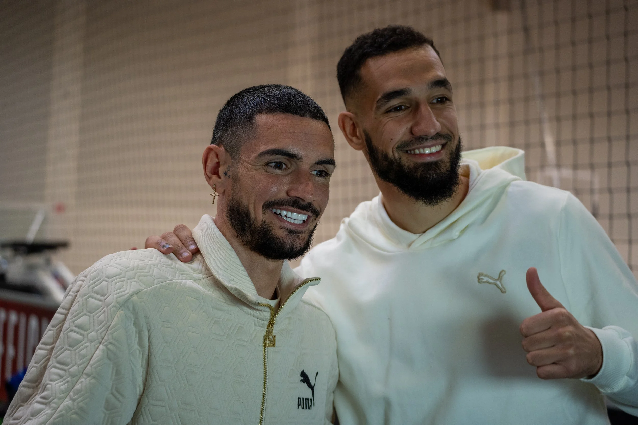 Deux hommes souriants prenant une photo ensemble, l'un avec un sweatshirt blanc avec un logo Puma et l'autre avec un sweat blanc avec un logo Puma, dans un lieu intérieur