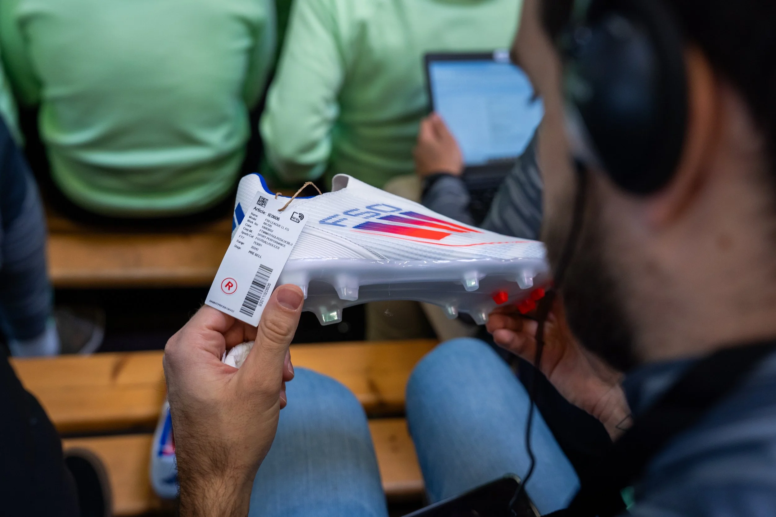 Une personne porte un casque et tient une chaussure de sport neuve blanche avec des détails rouges et bleus, probablement une chaussure de football, dans un gymnase ou une salle d'attente.