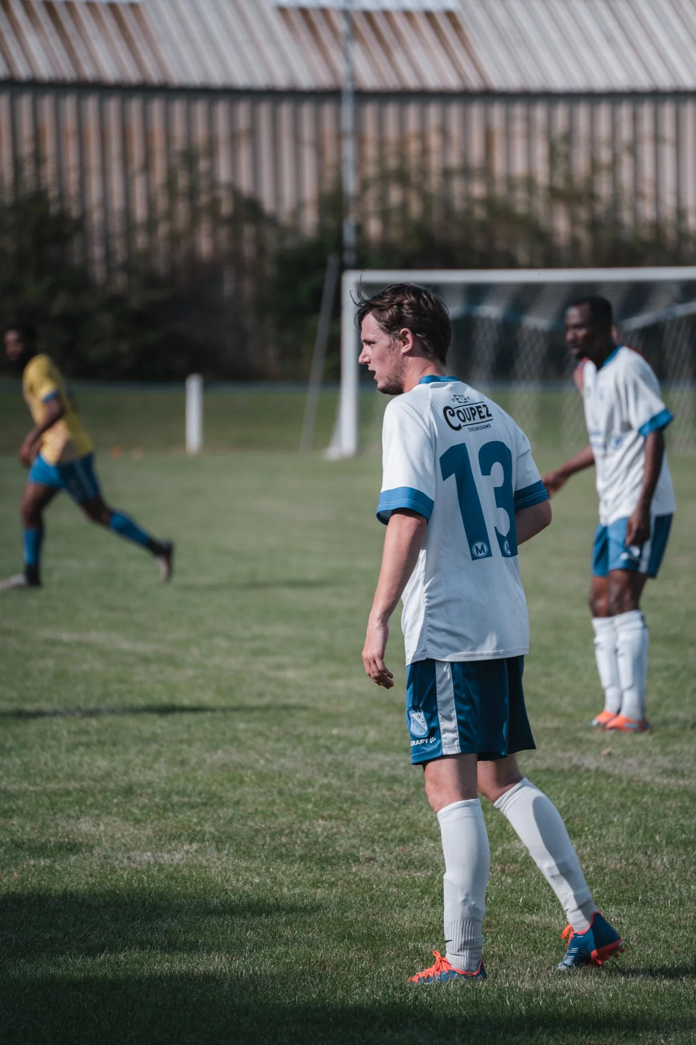 Joueur de football en blanc et bleu sur un terrains de sport, regardant vers le sol avec d'autres joueurs en arrière-plan.